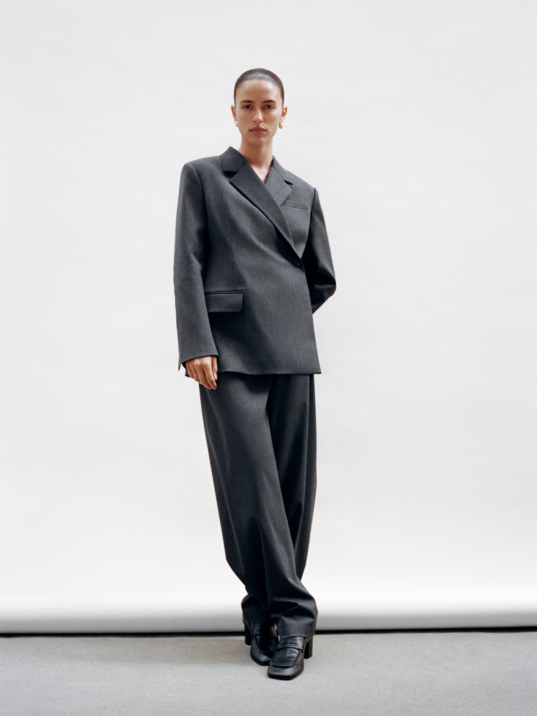 Größeres Bild anzeigen: Ein dunkelgrauer Anzug wird aus der Vorderansicht präsentiert, mit einem übergroßen zweireihigen Blazer mit breiten Kerbrevers, Pattentaschen und einem asymmetrischen Überkreuzverschluss. Dazu werden passende weite Hosen und schwarze Loafer kombiniert.