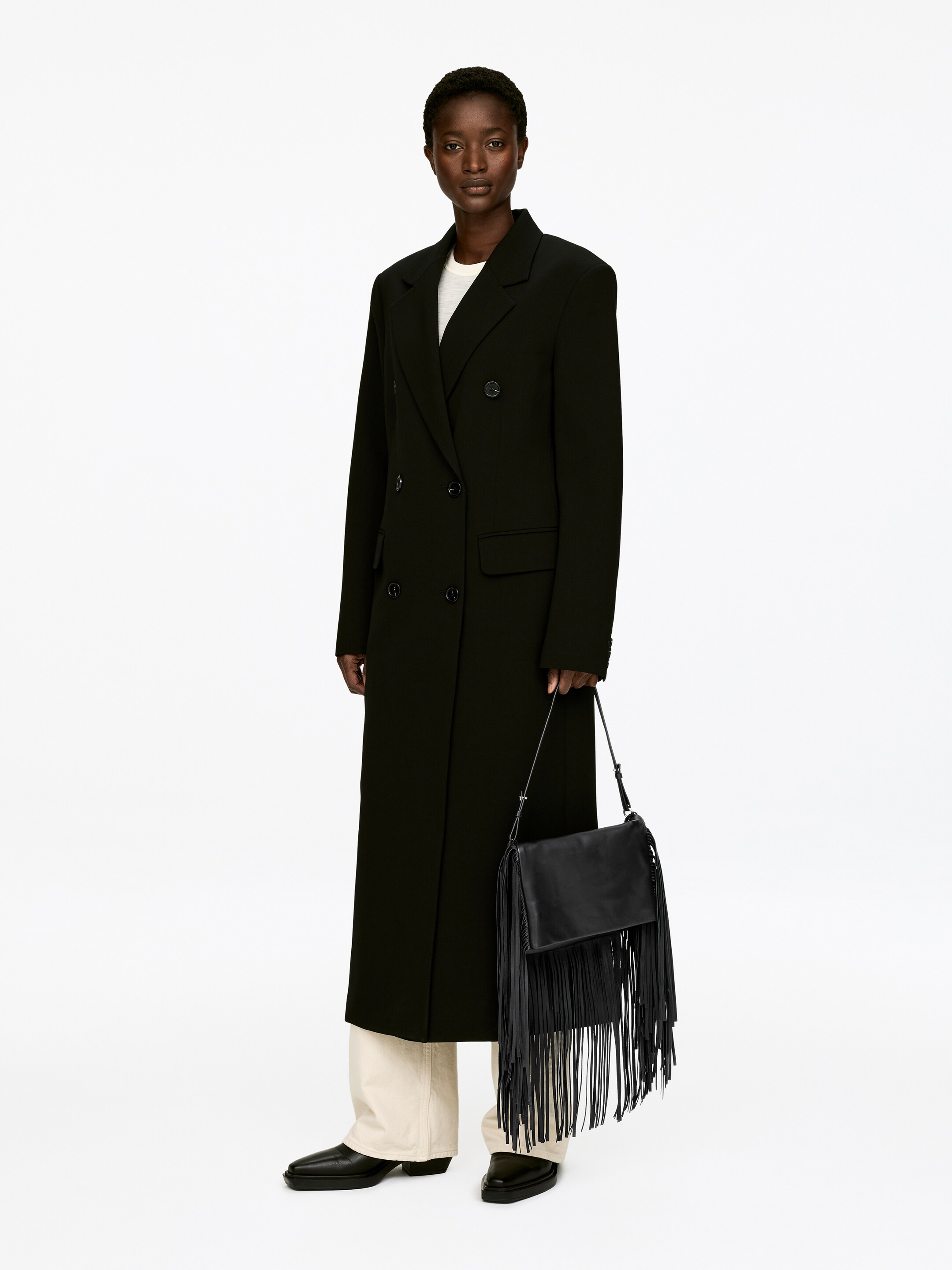 View larger image: A black double-breasted long coat is featuring notch lapels, structured shoulders, and two flap pockets, with four buttons on the front closure. It is facing slightly to the right, showcasing its mid-calf length over light-colored trousers and a visible light-colored top.