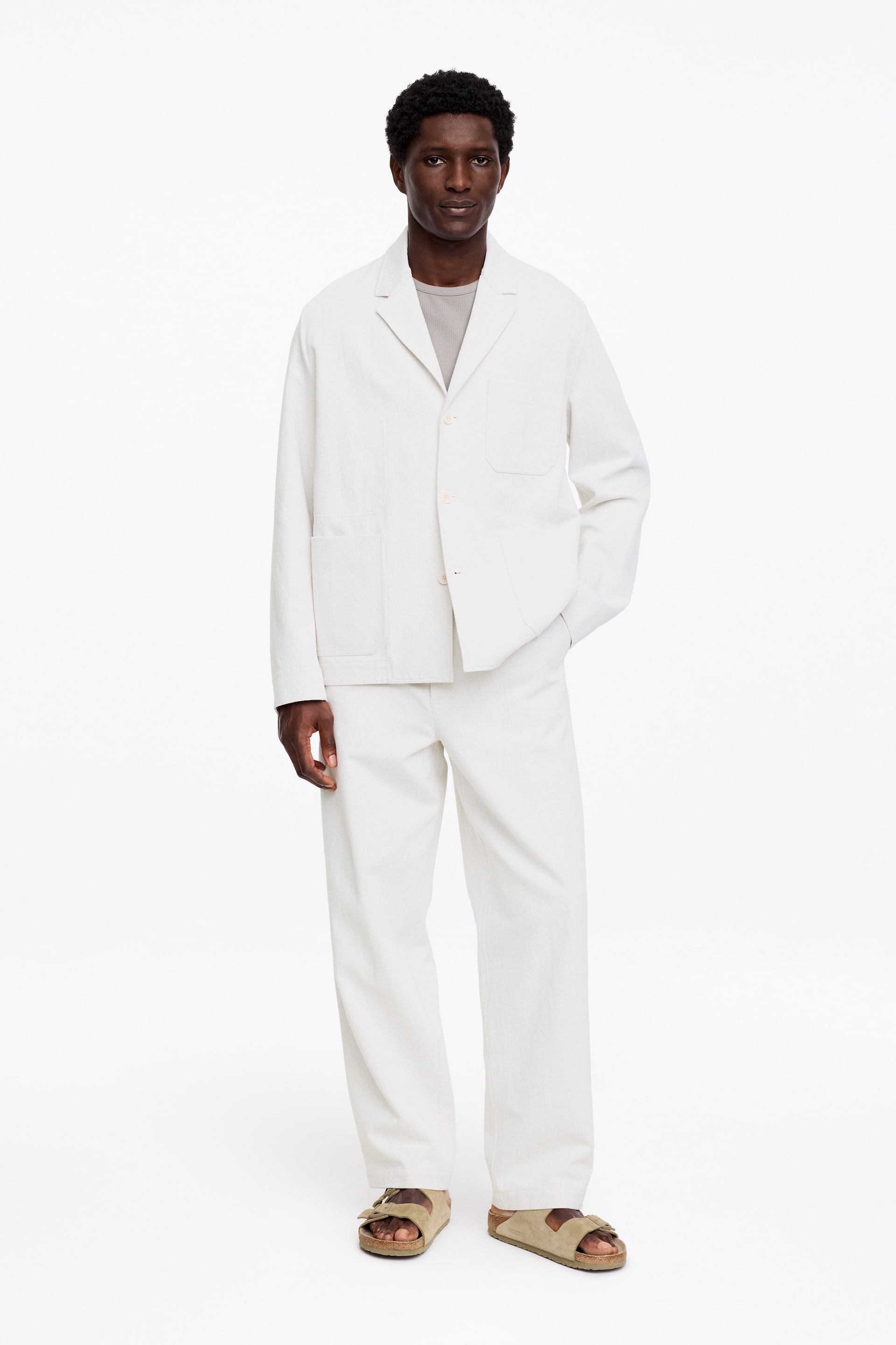 Grotere afbeelding bekijken: Een witte werkjas met inkeping revers en twee grote opgestikte zakken wordt gedragen over een lichtgrijs t-shirt, gecombineerd met bijpassende witte rechte broek. De volledige outfit is naar voren gericht, toont een ontspannen pasvorm en is gestyled met lichtbruine sandalen.