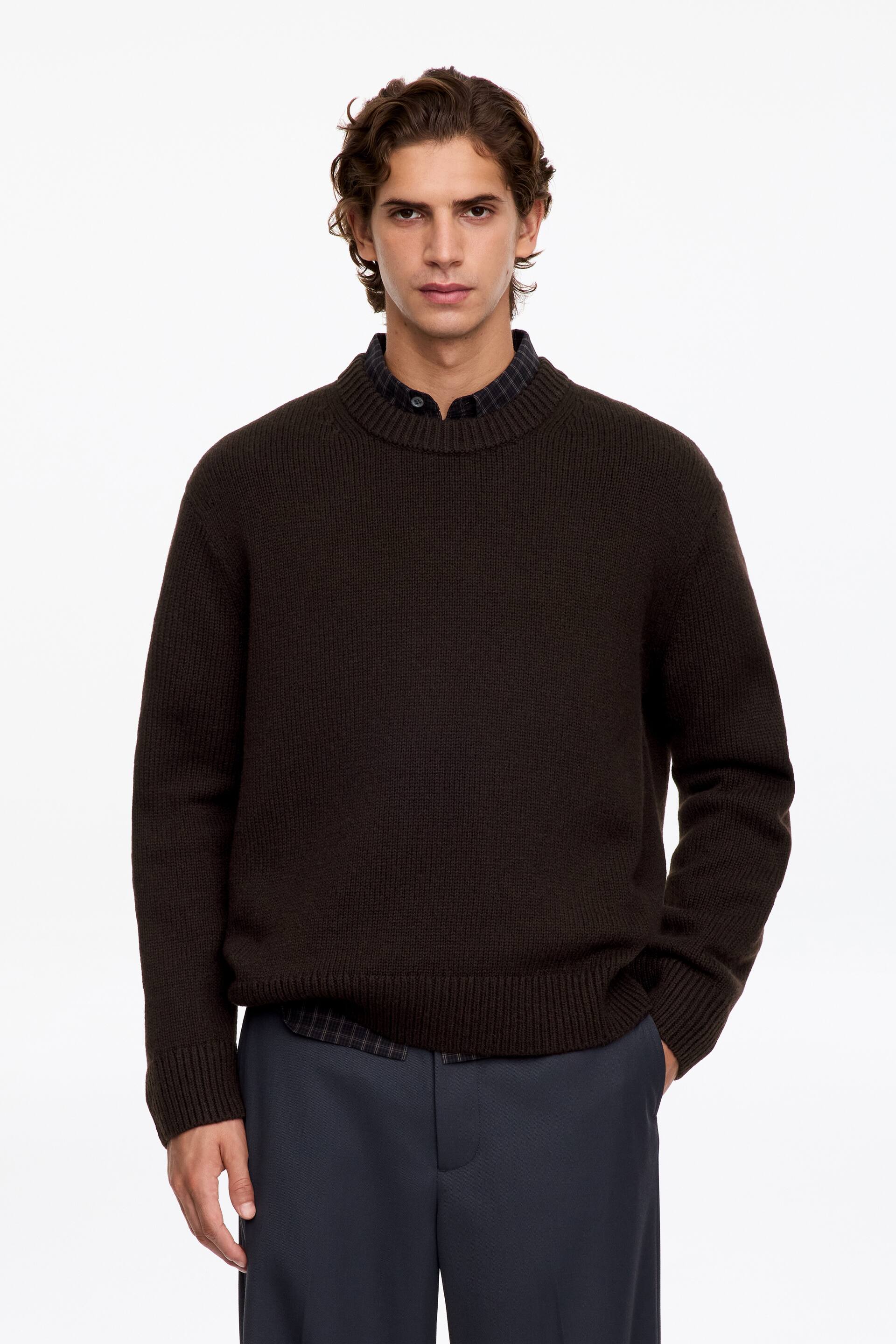 Ver imagen más grande: El modelo lleva puesto un jersey de cuello redondo marrón oscuro con cuello, puños y bajo de canalé grueso. El jersey se combina con una camisa oscura de cuello con estampado de cuadros y un pantalón oscuro.