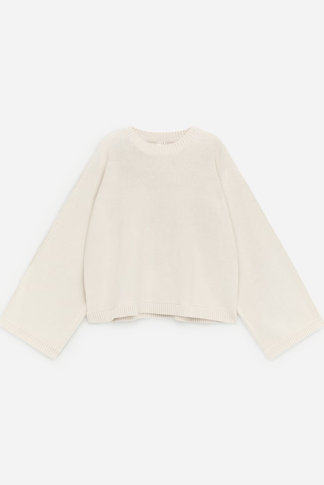 Un jersey de punto color crema se presenta de FRENTE, con un cuello redondo acanalado y ribetes acanalados en las mangas anchas, cortas y en el dobladillo. Se aprecia un pequeño detalle de aberturas a lo largo de la parte superior de las mangas. - 1