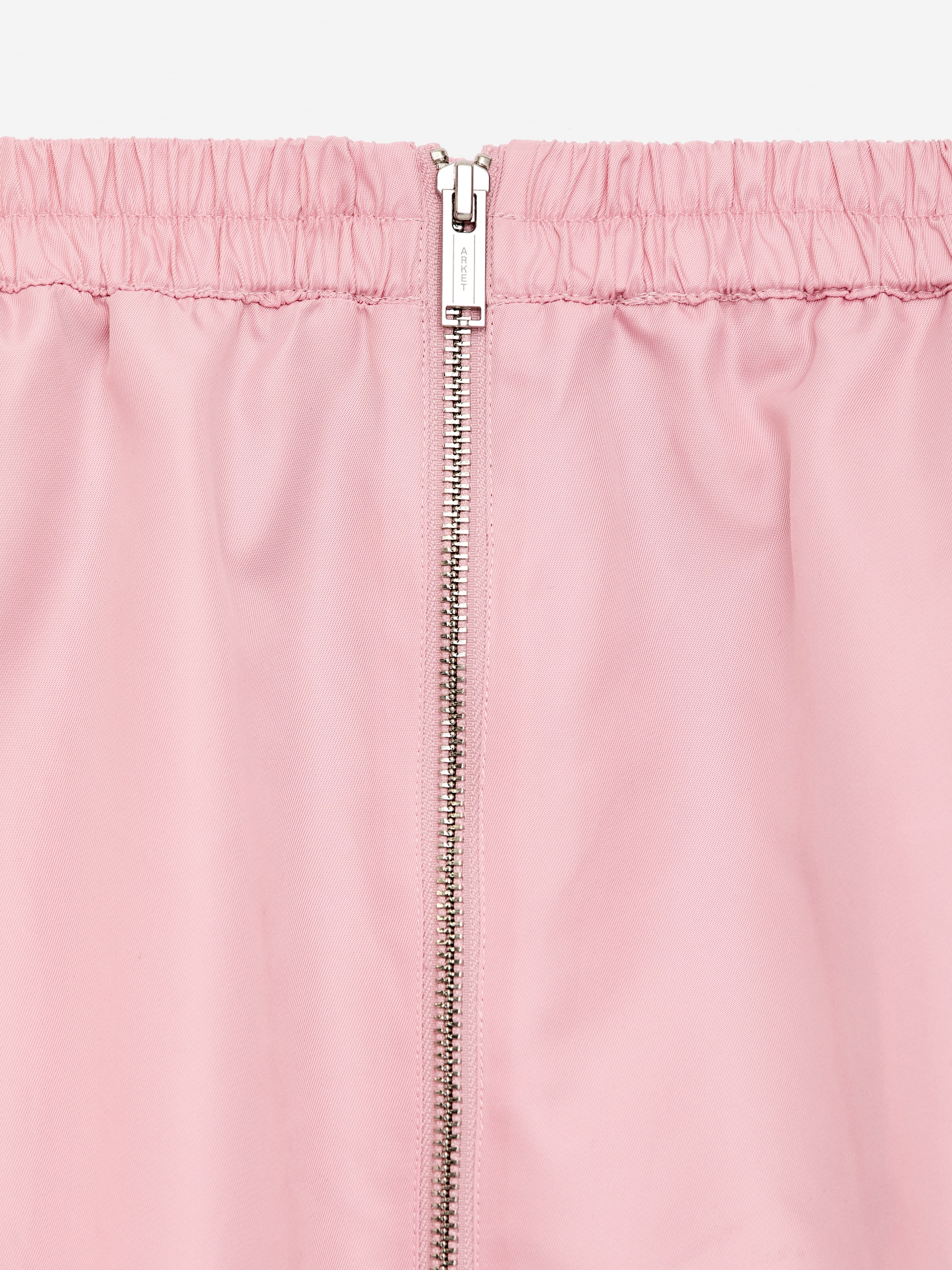 View larger image: A pink midi skirt is featuring an elasticated waistband and a metallic two-way zipper running vertically down its front. A single zipper pull, embossed with 'ARKET', is visible at the top of the zipper.