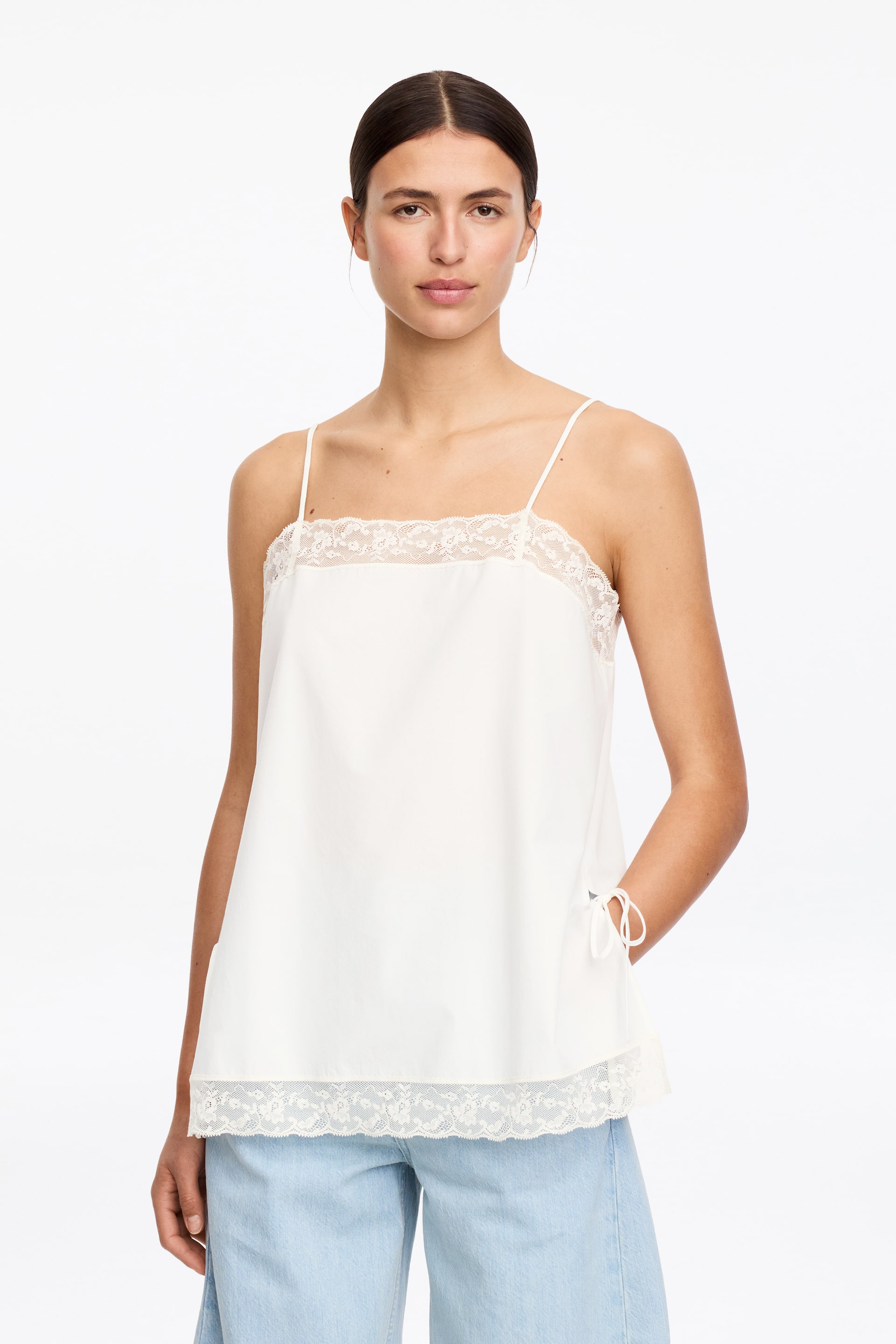 Grotere afbeelding bekijken: Een witte camisole top met dunne spaghettibandjes en een rechte halslijn met kantafwerking. De top heeft een ontspannen pasvorm, een rechte zoom met een bredere kanten rand, en decoratieve strikjes zijn zichtbaar aan de rechterkant.