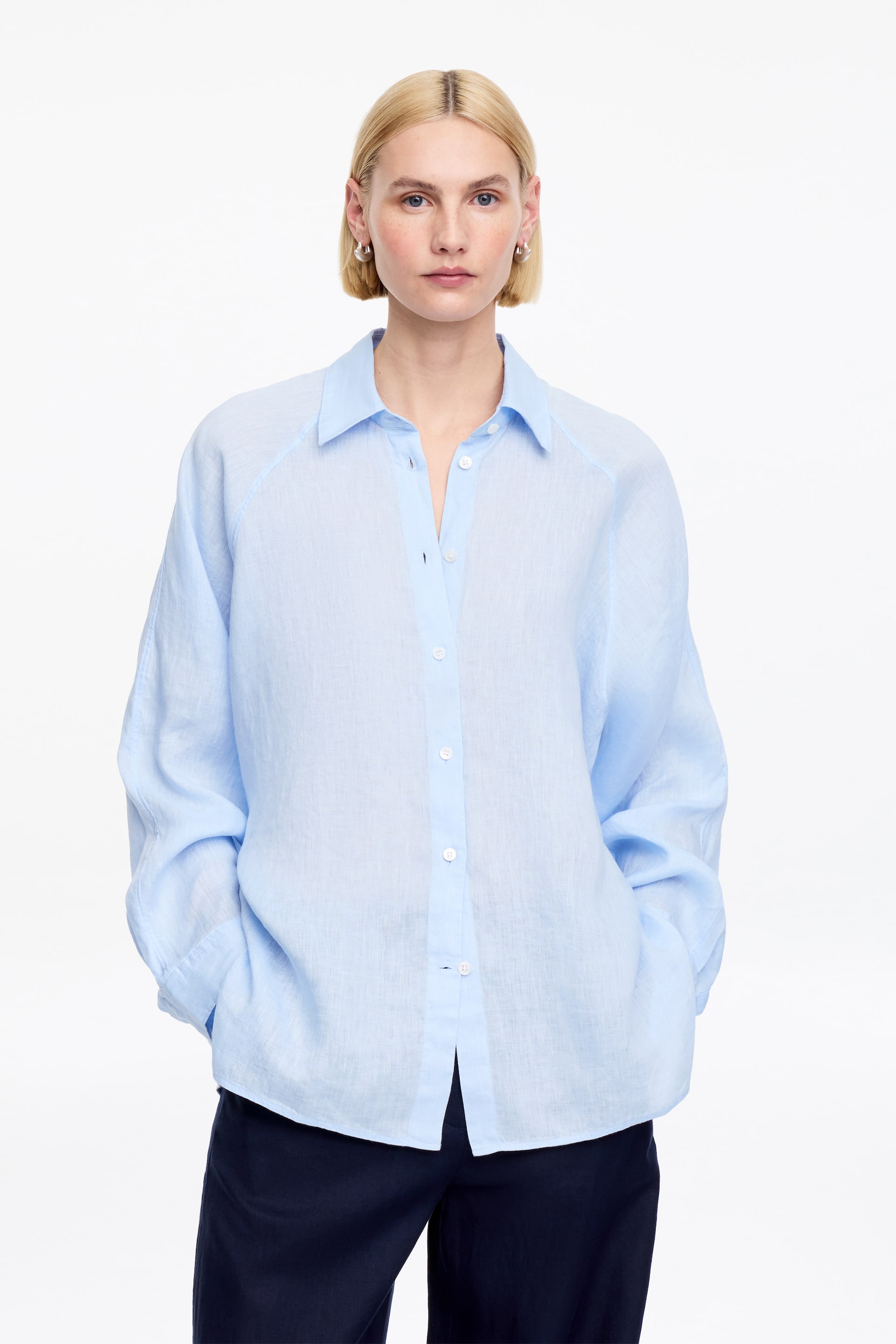 Agrandir l'image: Une chemise en lin bleu clair avec une coupe ample, des manches raglan et une fermeture boutonnée est portée par le mannequin. Le mannequin se tient debout avec les mains dans les poches d'un pantalon bleu foncé.