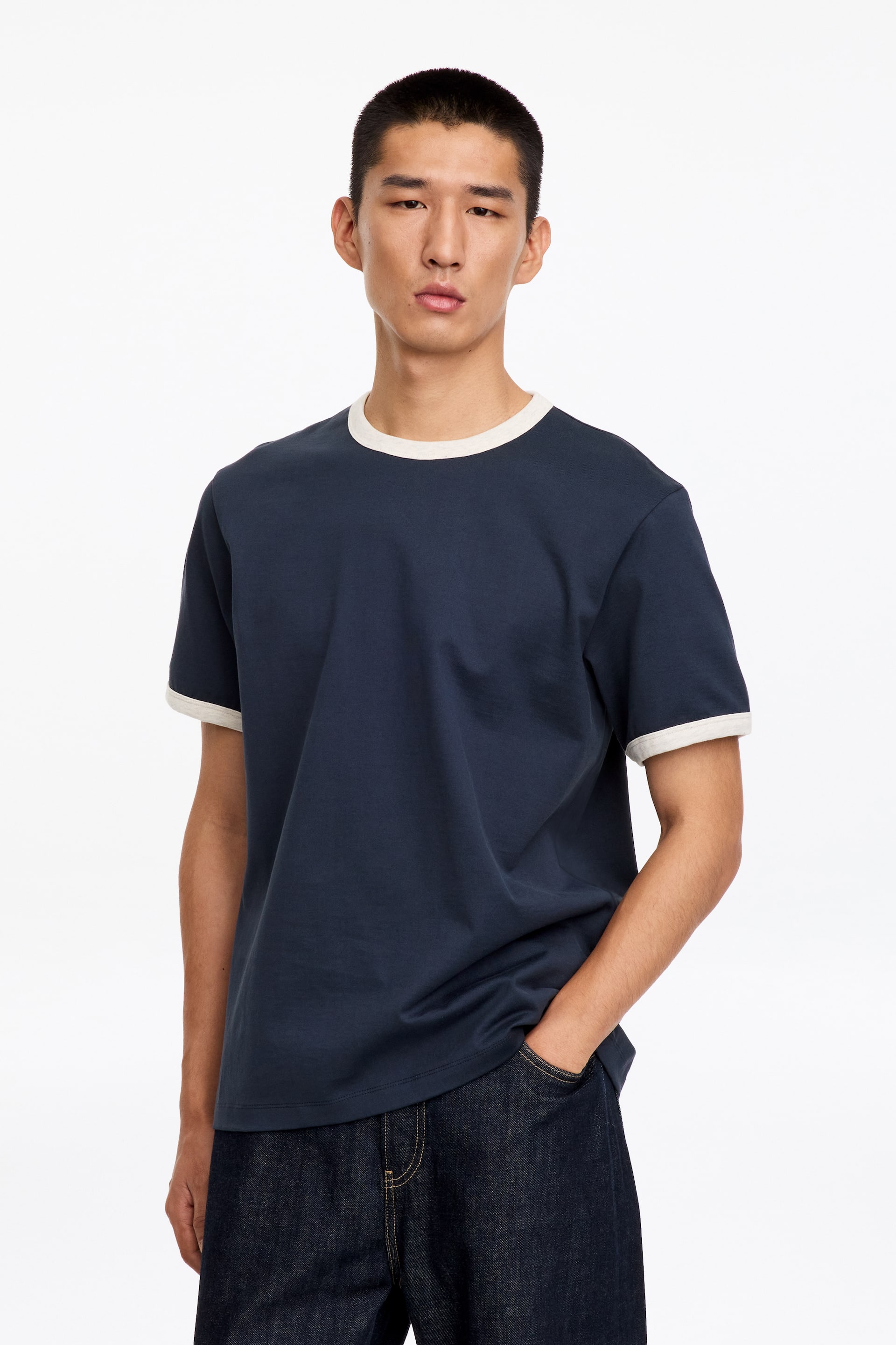 Ver imagen más grande: La modelo lleva puesta una camiseta azul oscuro con ribetes en contraste de color blanco roto en el cuello redondo y los puños de las mangas. La modelo está de pie con una mano metida en el bolsillo de unos vaqueros de denim oscuro.
