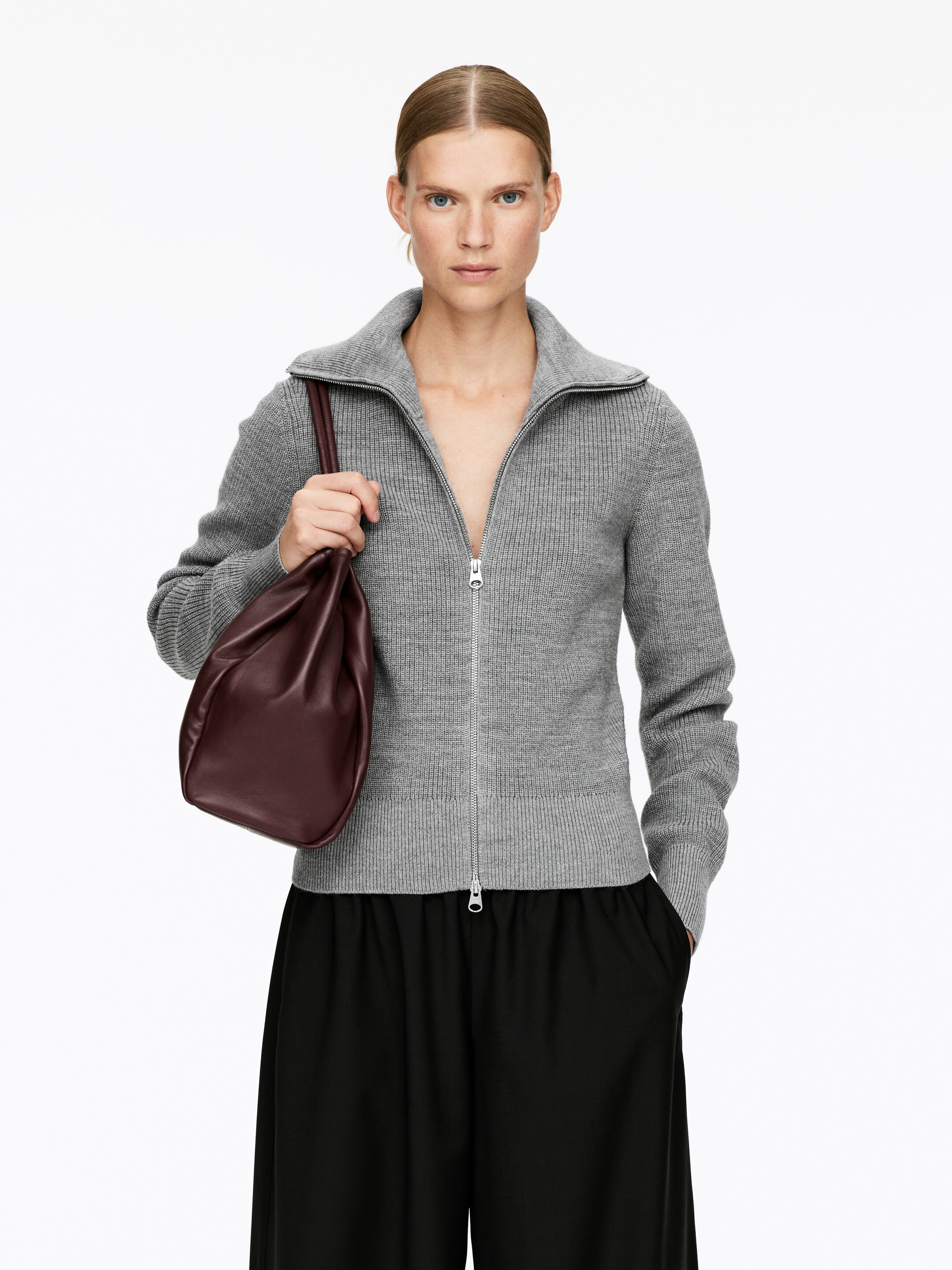 Vis større bilde: En grå ribbestrikket genser med full glidelås presenteres forfra, med en ståkrage, en synlig metallglidelås og ribbestrikkede mansjetter og kant. En mørkerød, myk, slouchy veske holdes av brukeren mot genseren.