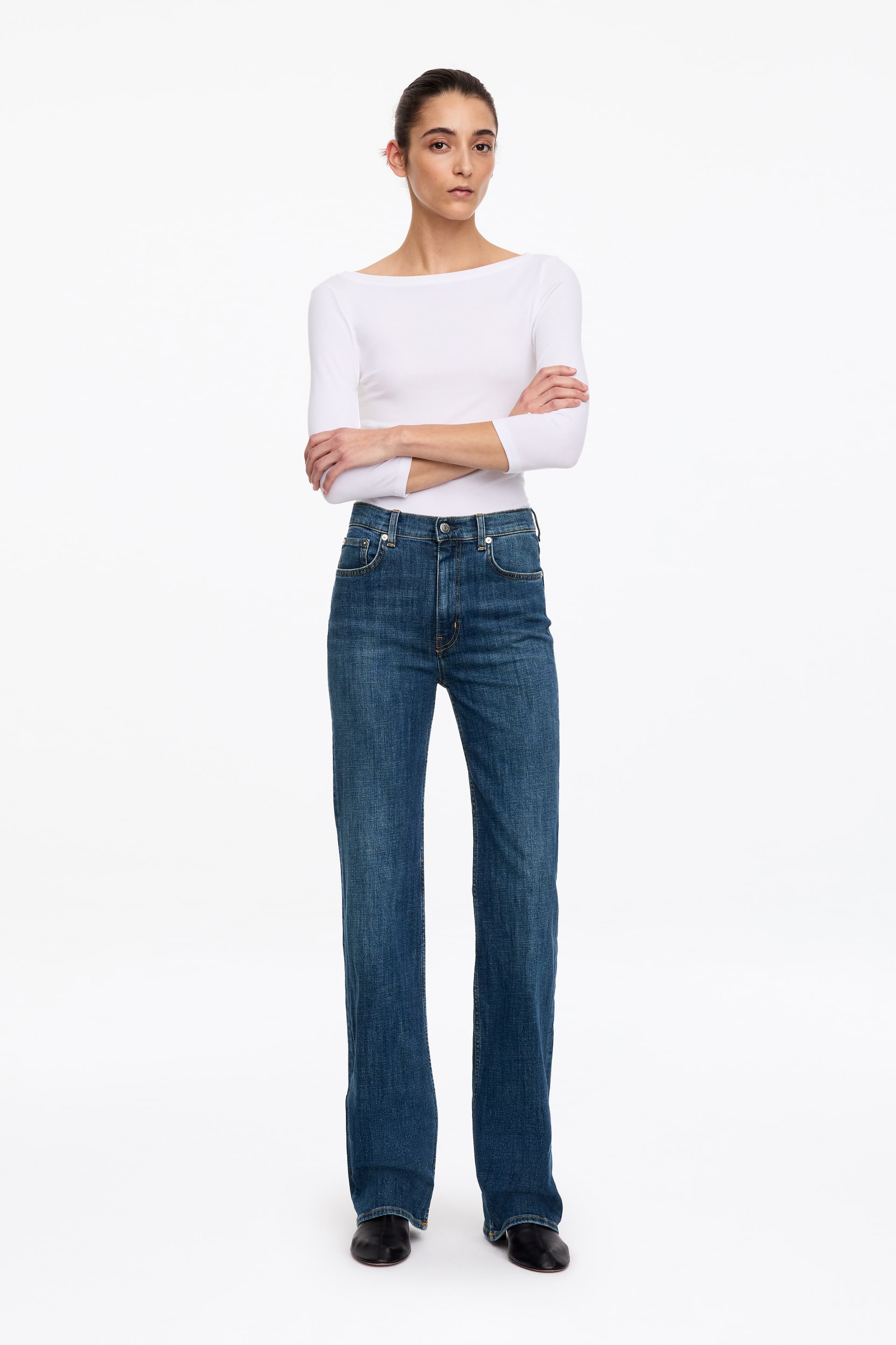 Agrandir l'image: Un jean en denim bleu, doté d’une taille haute, d’une coupe slim et de jambes bootcut, est porté par le mannequin. Le jean est associé à un haut blanc à manches longues rentré à l’intérieur et à des chaussures noires.