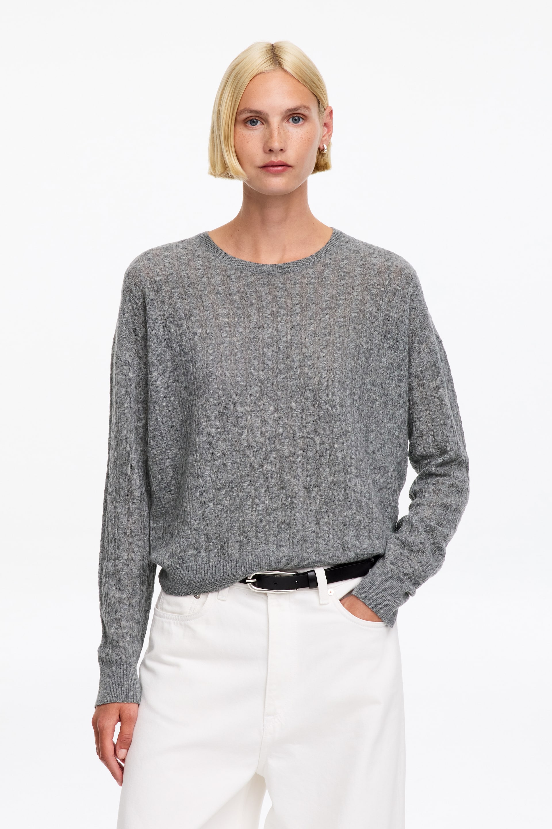 Ver imagem maior: A modelo está a usar uma camisola cinzenta leve de malha canelada, com decote redondo e mangas compridas, de corte descontraído. A camisola está combinada com calças brancas e um cinto preto, e uma das mãos da modelo está pousada num bolso das calças.