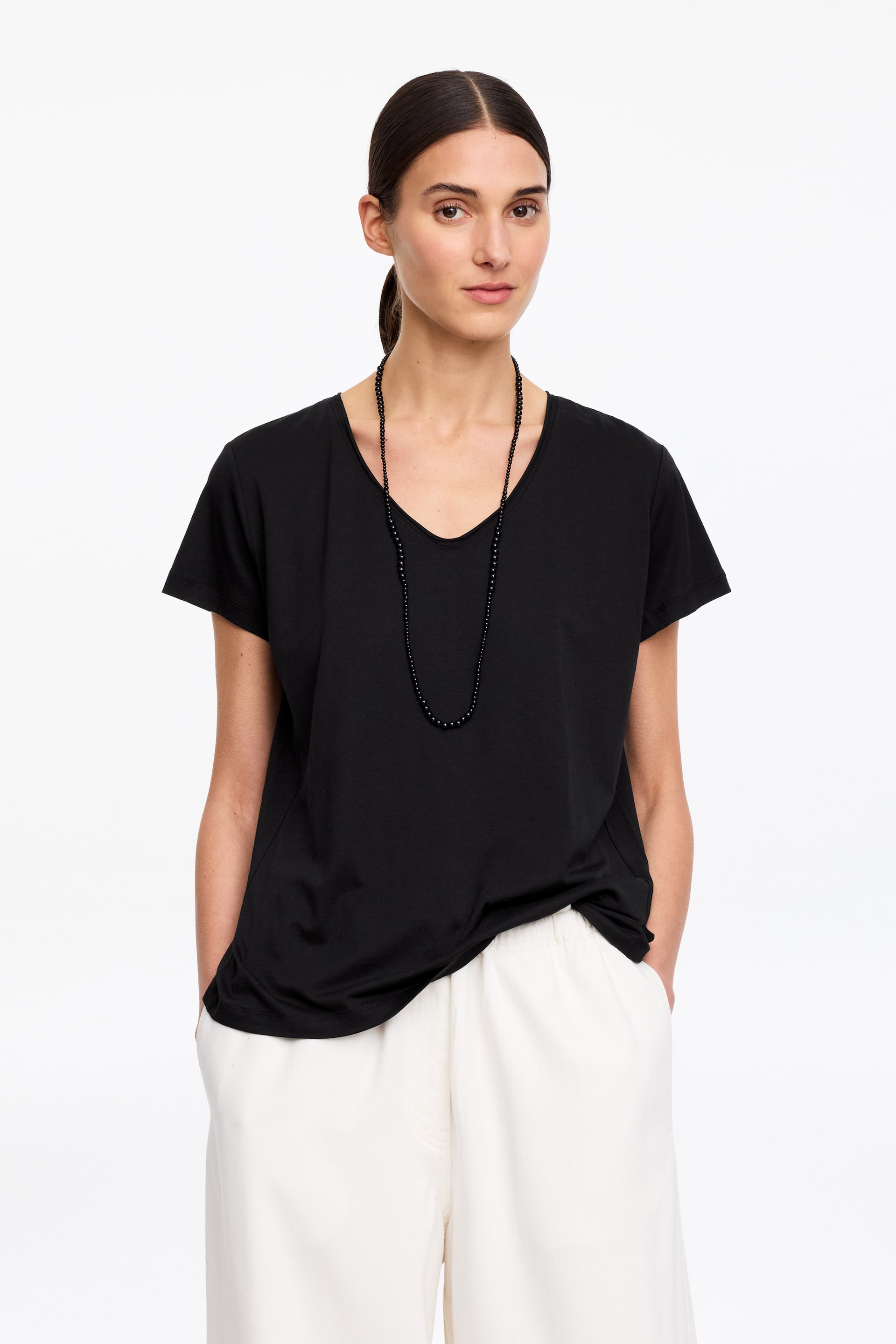 Ingrandisci l'immagine: La modella indossa una maglietta nera a maniche corte. Presenta uno scollo a V e cuciture laterali curve che si spostano verso il davanti. Una lunga collana nera con perline è drappeggiata sulla maglietta.