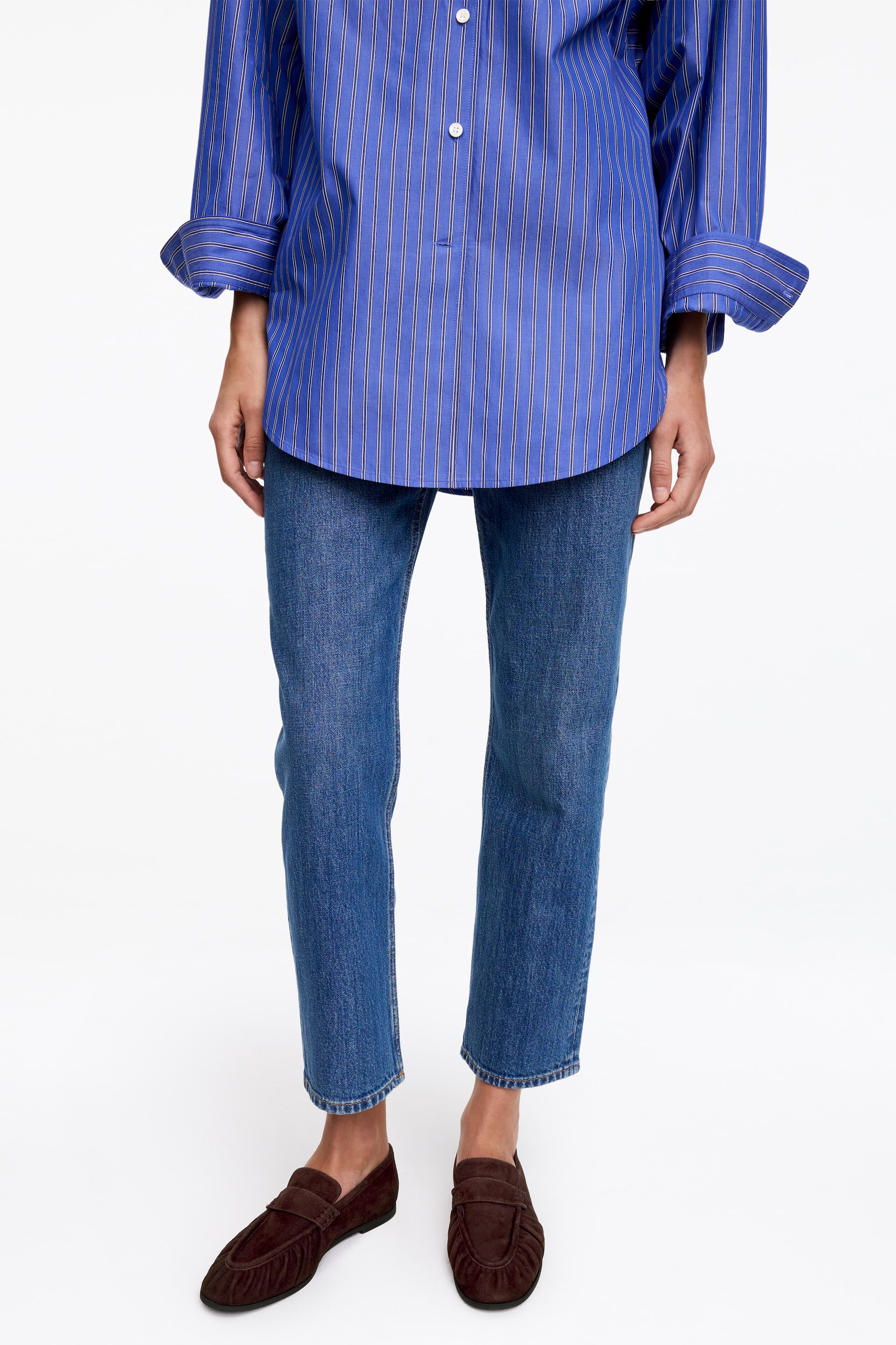 Vis større bilde: En blå skjorte med tynne, vertikale hvite striper, delvis knappestolpe foran med hvite knapper, og lange ermer med oppbrettede mansjetter; den har en avslappet passform og faller løst over blå jeans.