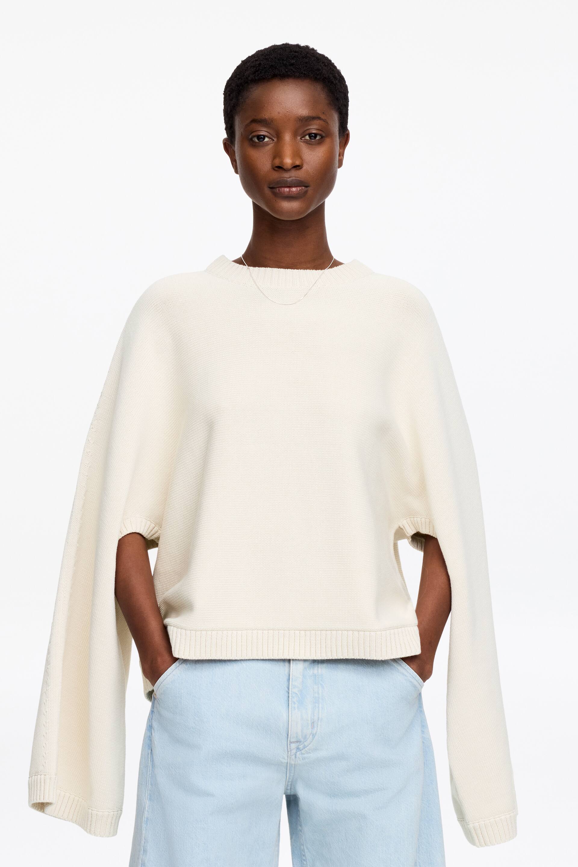 Grotere afbeelding bekijken: Een gebroken witte trui wordt frontaal gepresenteerd, met een ronde hals en een uniek ontwerp met open zijkanten van de mouwen. De trui heeft een geribde zoom en geribde boorden aan de lange, gespleten mouwen, met een dunne zilveren ketting die op de halslijn rust.