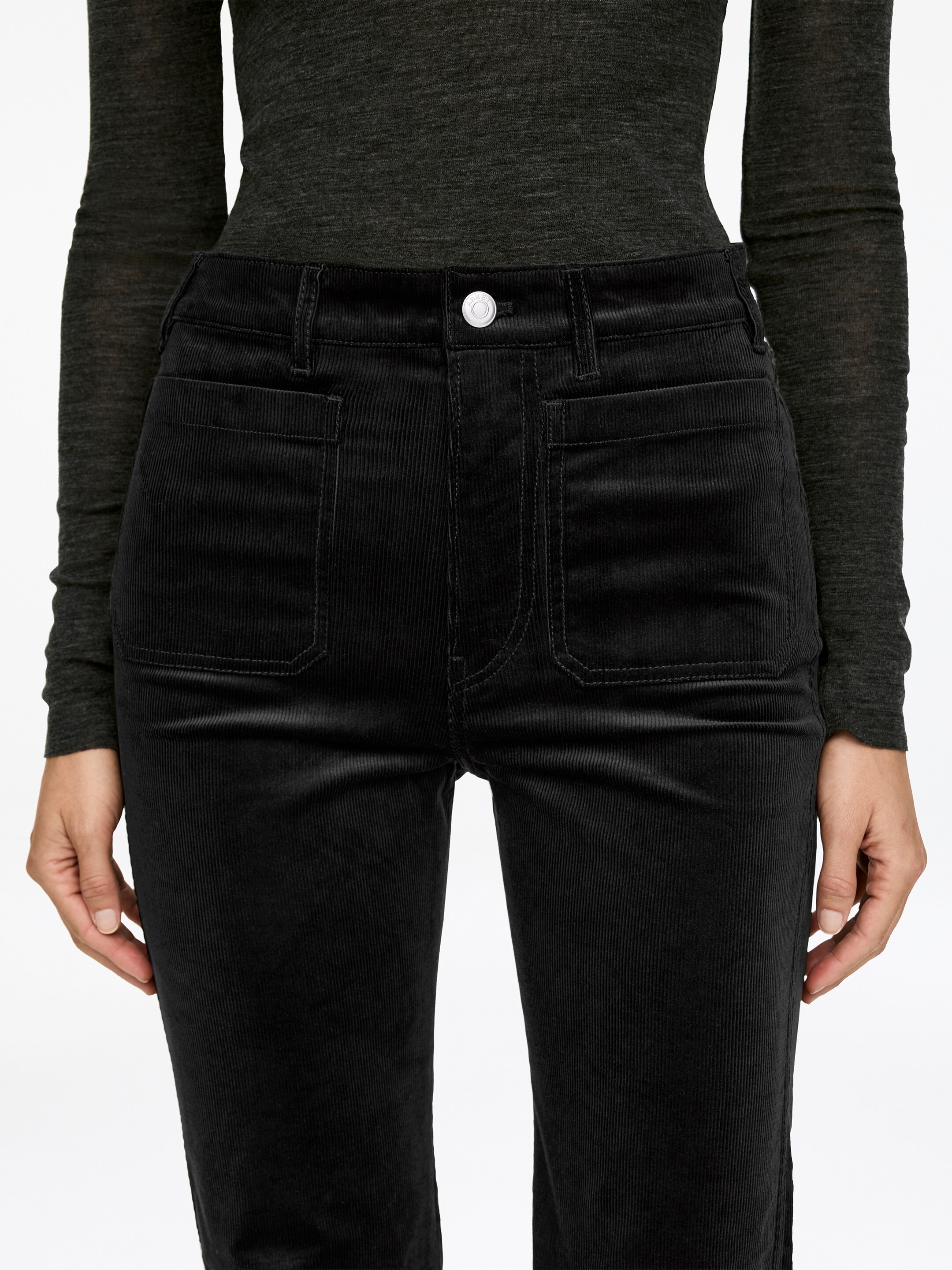 Ingrandisci l'immagine: Pantaloni in velluto a coste nero, caratterizzati da una vita alta, tasche frontali a toppa e solchi verticali visibili, vengono indossati dalla modella. Questi pantaloni a vestibilità slim hanno gambe svasate e sono abbinati a un top scuro a maniche lunghe infilato nei pantaloni, mentre le mani della modella riposano ai lati.