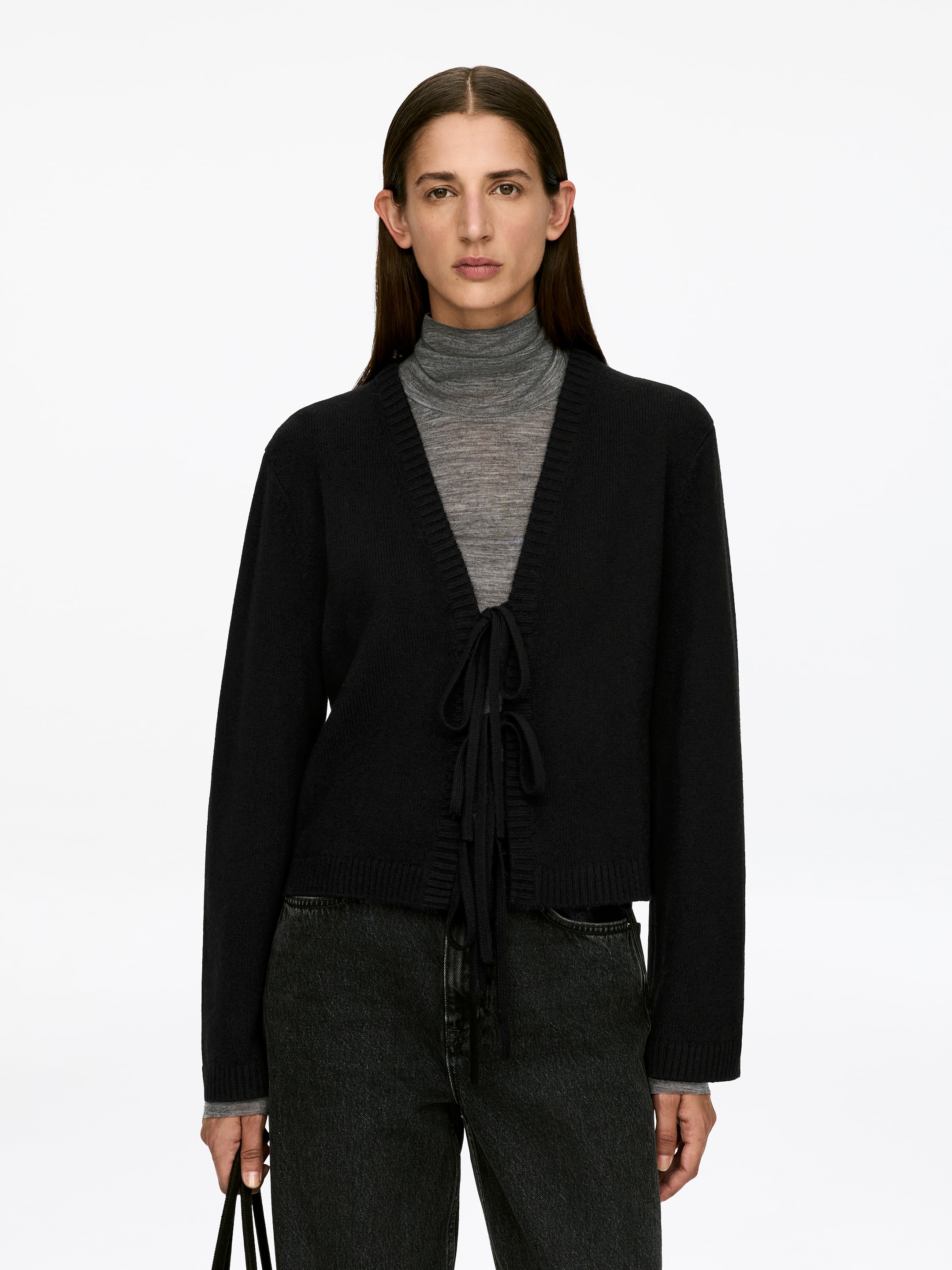 Vis større bilde: En svart cardigan i ren ull, med tre knyttede frontbånd og ribbestrikkede kanter, bæres av modellen. Den er lagd over en grå høyhalset genser og stylet med sorte jeans.
