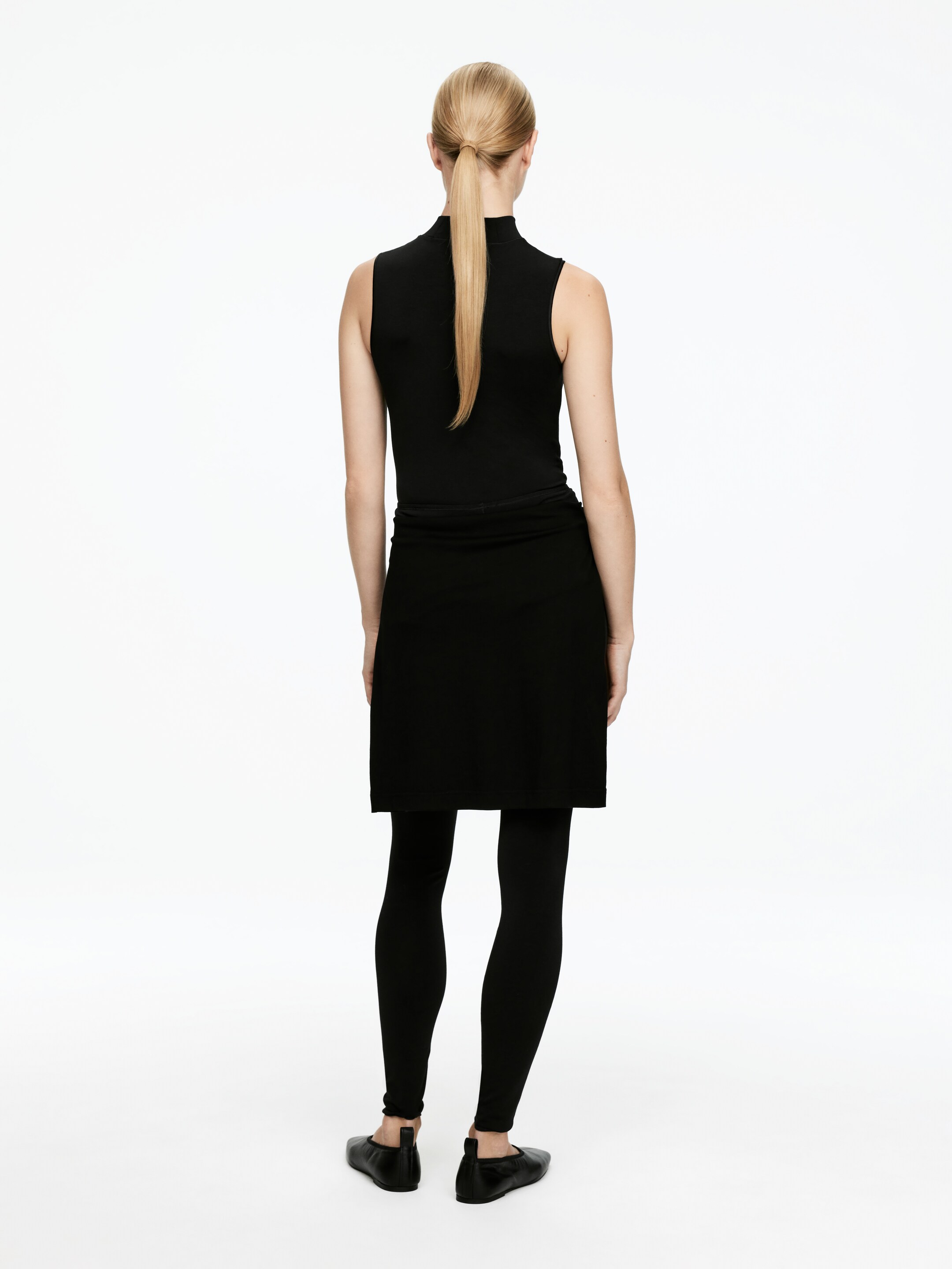 Agrandir l'image: Un haut noir sans manches à col montant est porté, associé à une jupe courte noire superposée à des leggings noirs ajustés, et des chaussures plates noires. L'ensemble est présenté de dos.