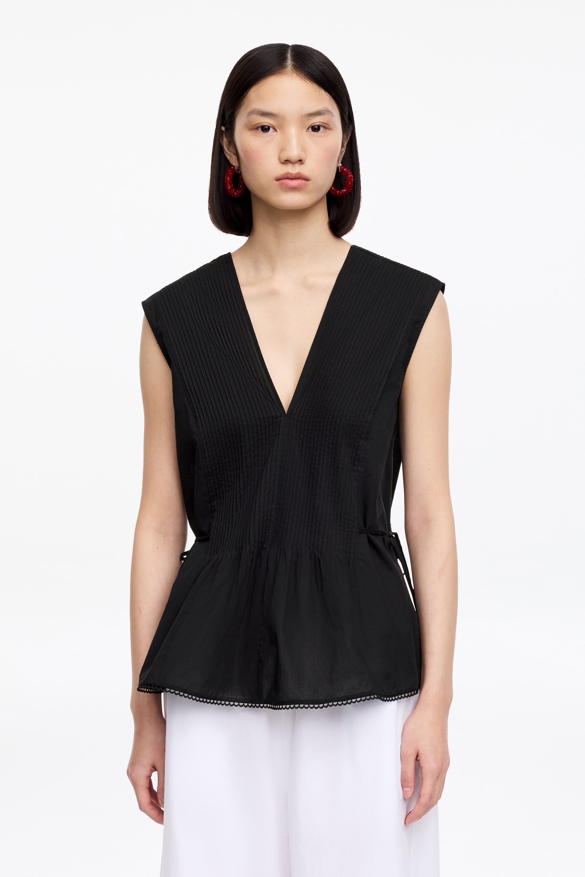 Ver imagem maior: Um top preto sem mangas apresenta um decote em V profundo e pregas no corpete, estendendo-se dos ombros até a metade do torso. É desenhado com um flare estilo peplum a partir da metade do torso, incluindo cordões ajustáveis nas laterais e um acabamento recortado ao longo da bainha inferior.