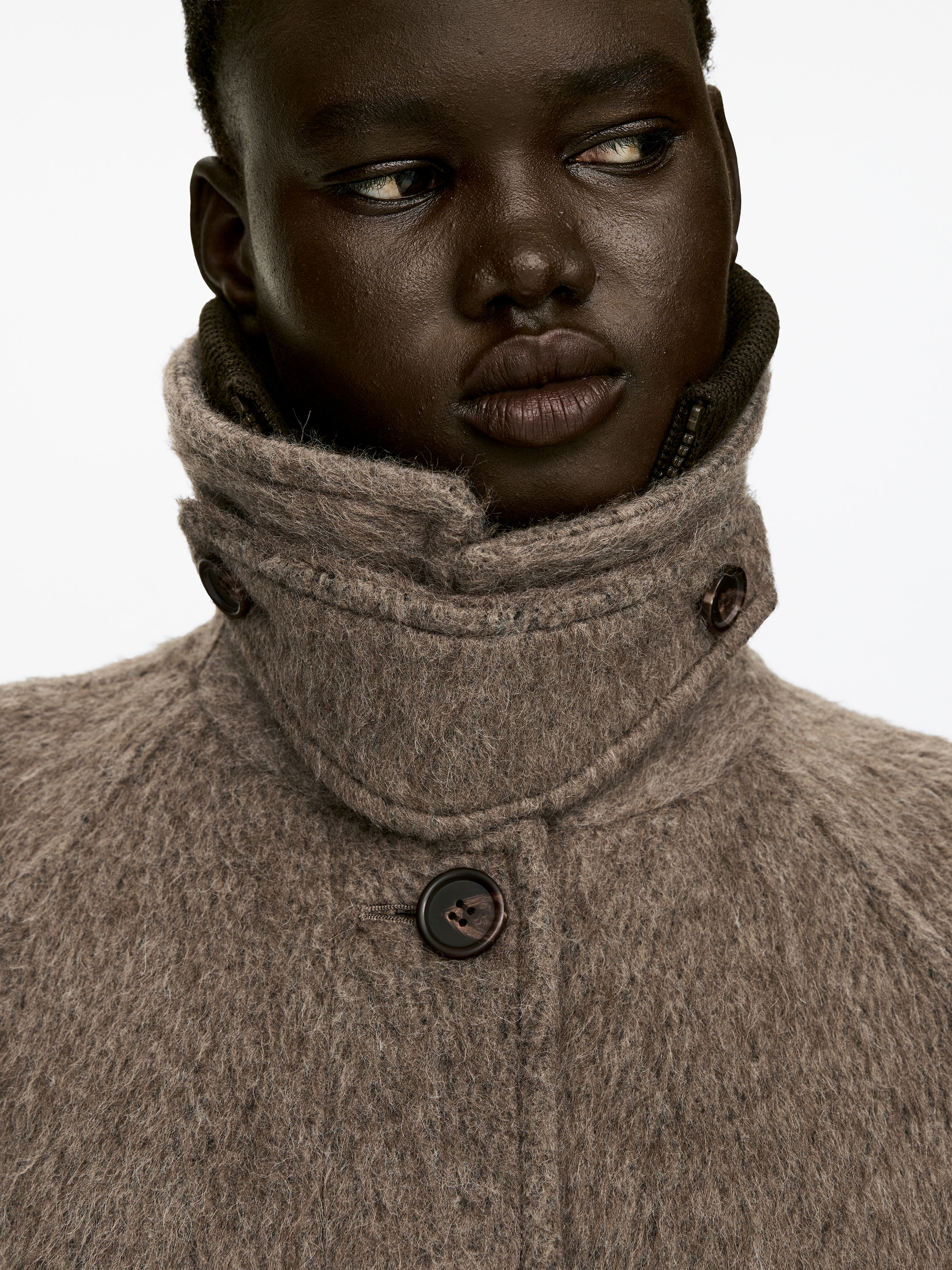 View larger image: A single-breasted coat in a textured, brownish-grey brushed wool and alpaca blend is being worn by the model. Its standing collar is buttoned, with an inner dark ribbed detail and zipper showing, while a front button is partially visible.