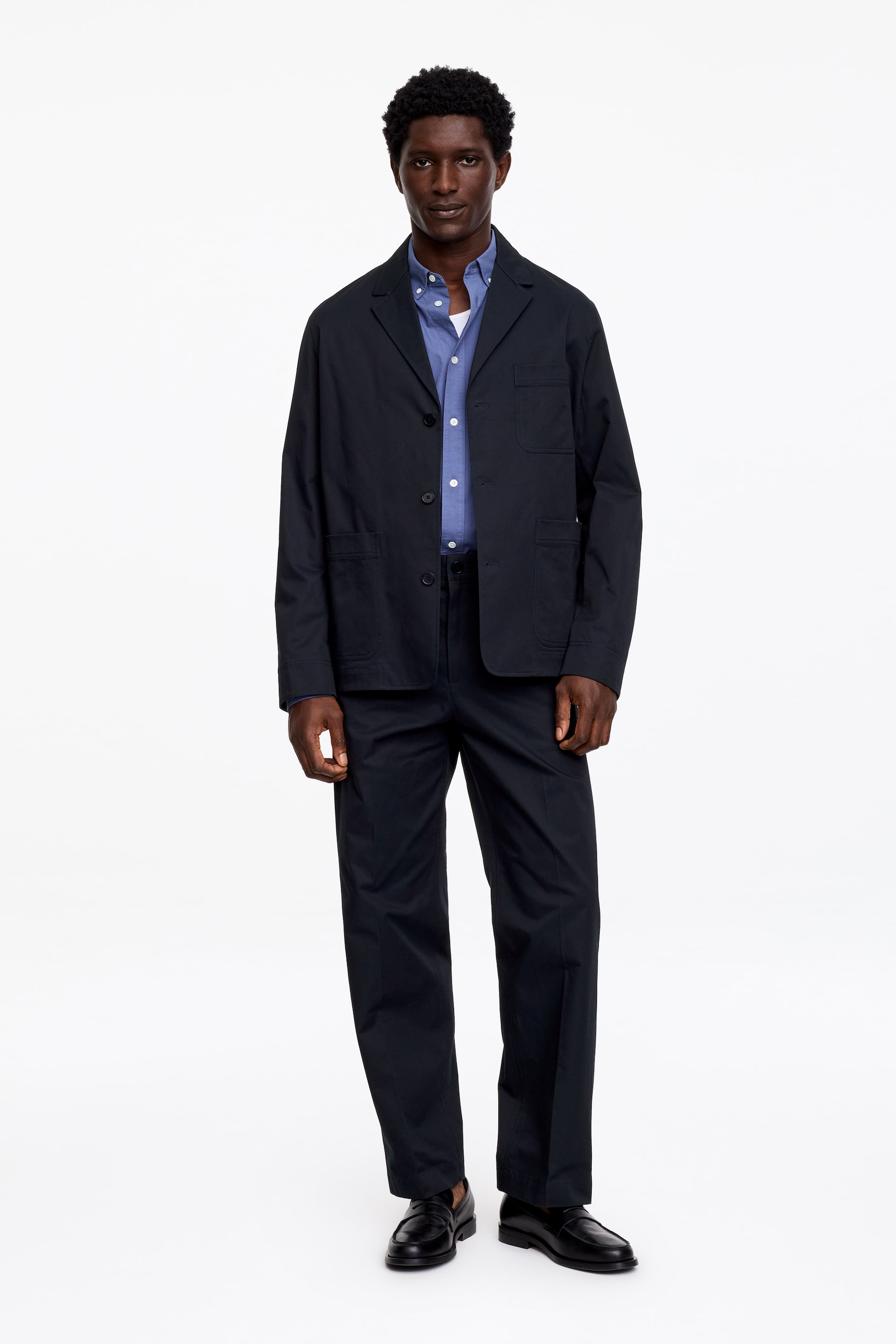 Visa större bild: En mörkblå avslappnad kostym, med en enkelknäppt kavaj med kavajslag och påsydda fickor, bärs öppen över en ljusblå skjorta med knappar. Den presenteras med matchande raka byxor och svarta loafers i läder.