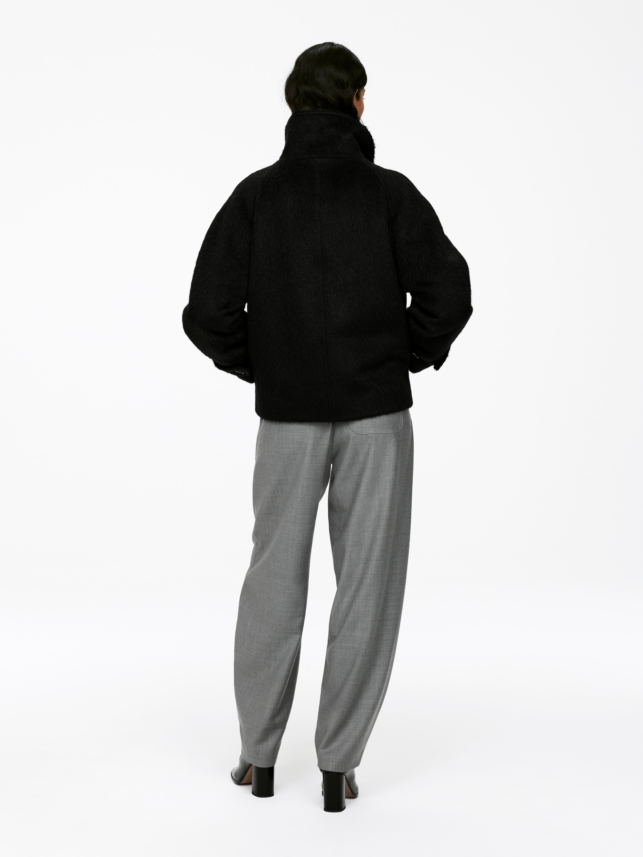 Agrandir l'image: Une veste noire, texturée et courte est vue de dos, avec un col montant et des manches larges à épaules tombantes. Elle est associée à un pantalon gris taille haute à coupe décontractée qui s'accumule légèrement aux chevilles, complété par des bottes à talons blocs noires.