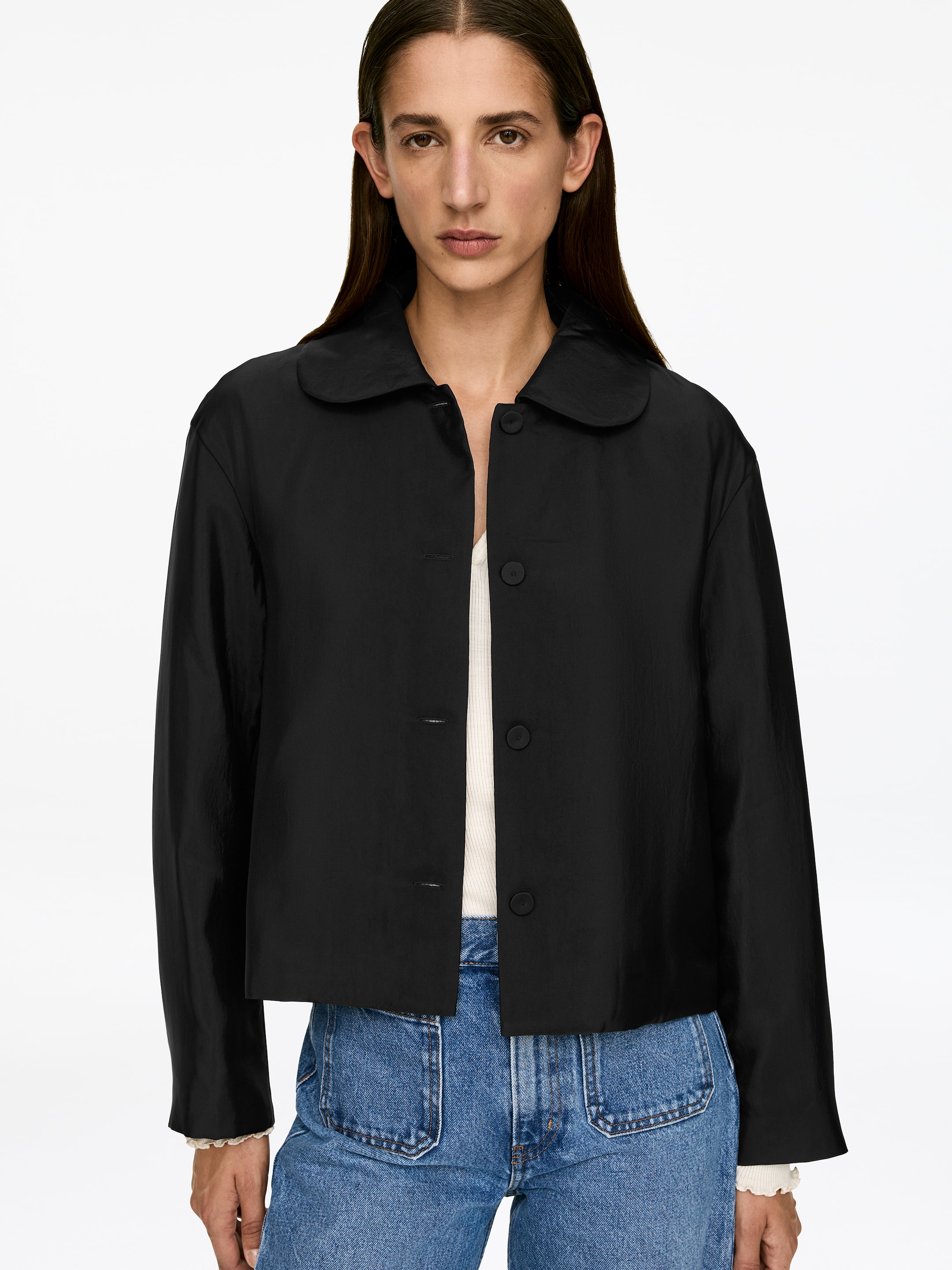 Größeres Bild ansehen: Ein schwarzes gepolstertes Overshirt mit einem abgerundeten Kragen und stoffbezogenen Knöpfen wird von dem Modell offen getragen. Darunter ist ein weißes geripptes Oberteil zu sehen, das mit blauen Jeans kombiniert wird.
