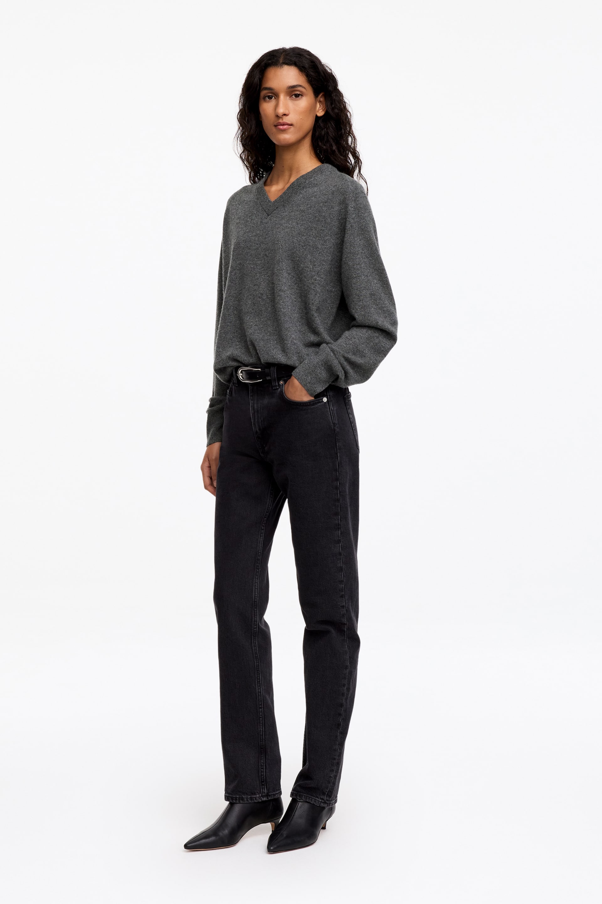 Visa större bild: En grå jumper med avslappnad passform och V-ringning bärs av modellen, med långa ärmar och ribbade muddar. Jumpern är stylad med framkanten löst instoppad i svarta jeans.