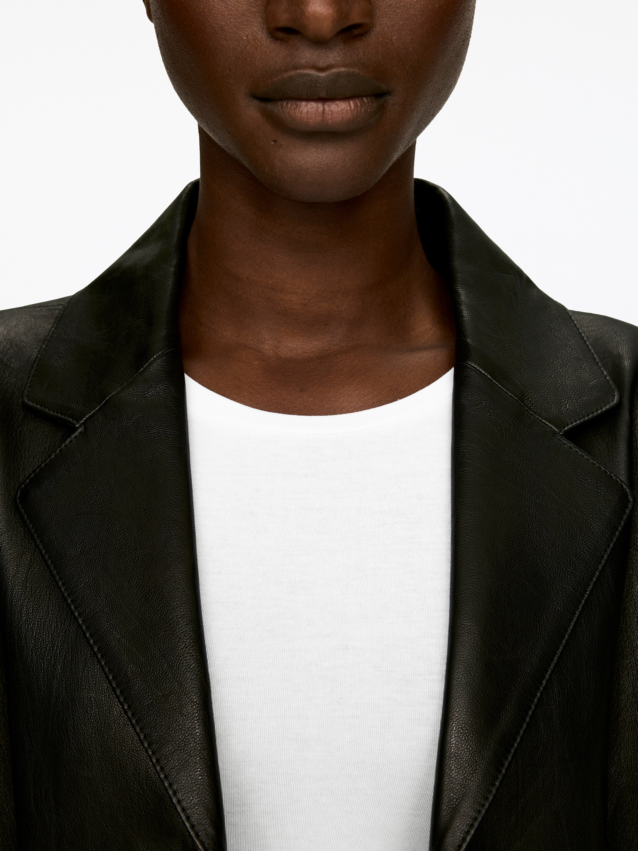 Agrandir l'image: Une veste en cuir noir est montrée de face, couvrant le haut du torse. Elle présente de larges revers crantés avec des coutures visibles le long de leurs bords et est portée ouverte sur un t-shirt à col rond blanc.