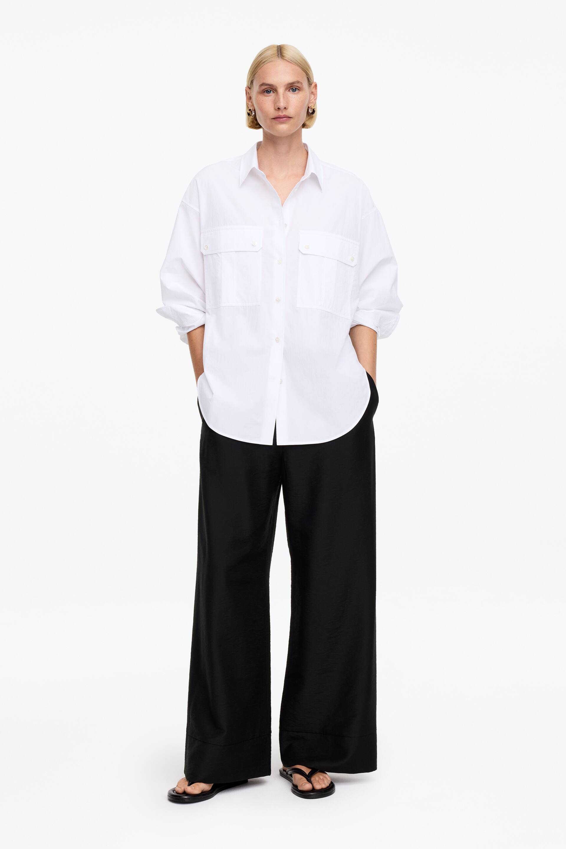 Zobacz większy obraz: Biała koszula zapinana na guziki z kołnierzykiem w szpic, dwoma kieszeniami na piersi z klapkami zapinanymi na guziki oraz podwiniętymi długimi rękawami. Prezentuje luźny krój w subtelnie teksturowanej tkaninie.