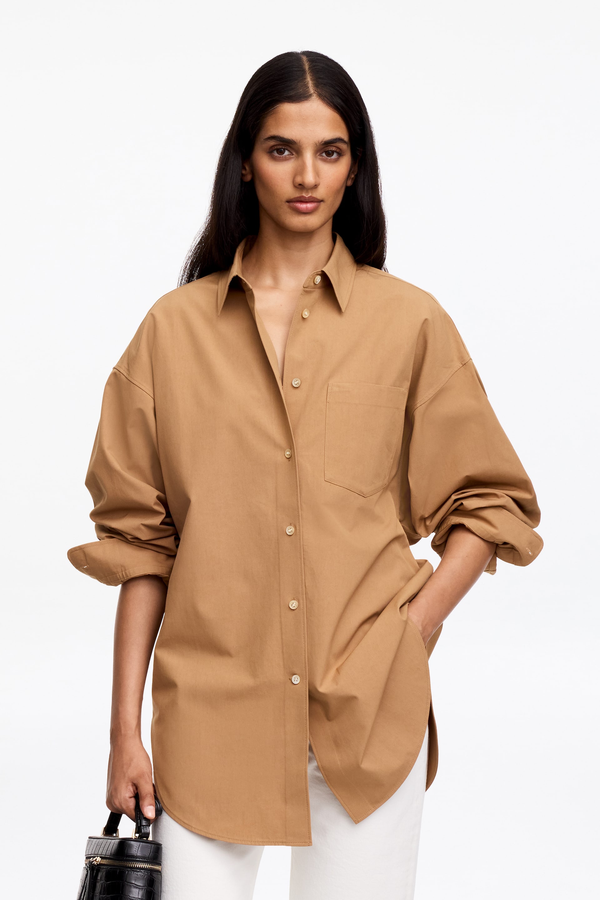 Agrandir l'image: Une chemise beige à boutons est portée, vue de face. Elle présente un col pointu et des boutons de couleur claire sur sa patte de boutonnage, avec une poche plaquée unique sur la poitrine gauche. Ses longues manches oversize sont retroussées, et une main repose dans une poche latérale, mettant en valeur une coupe allongée et décontractée avec un ourlet arrondi, portée non rentrée sur un pantalon de couleur claire.