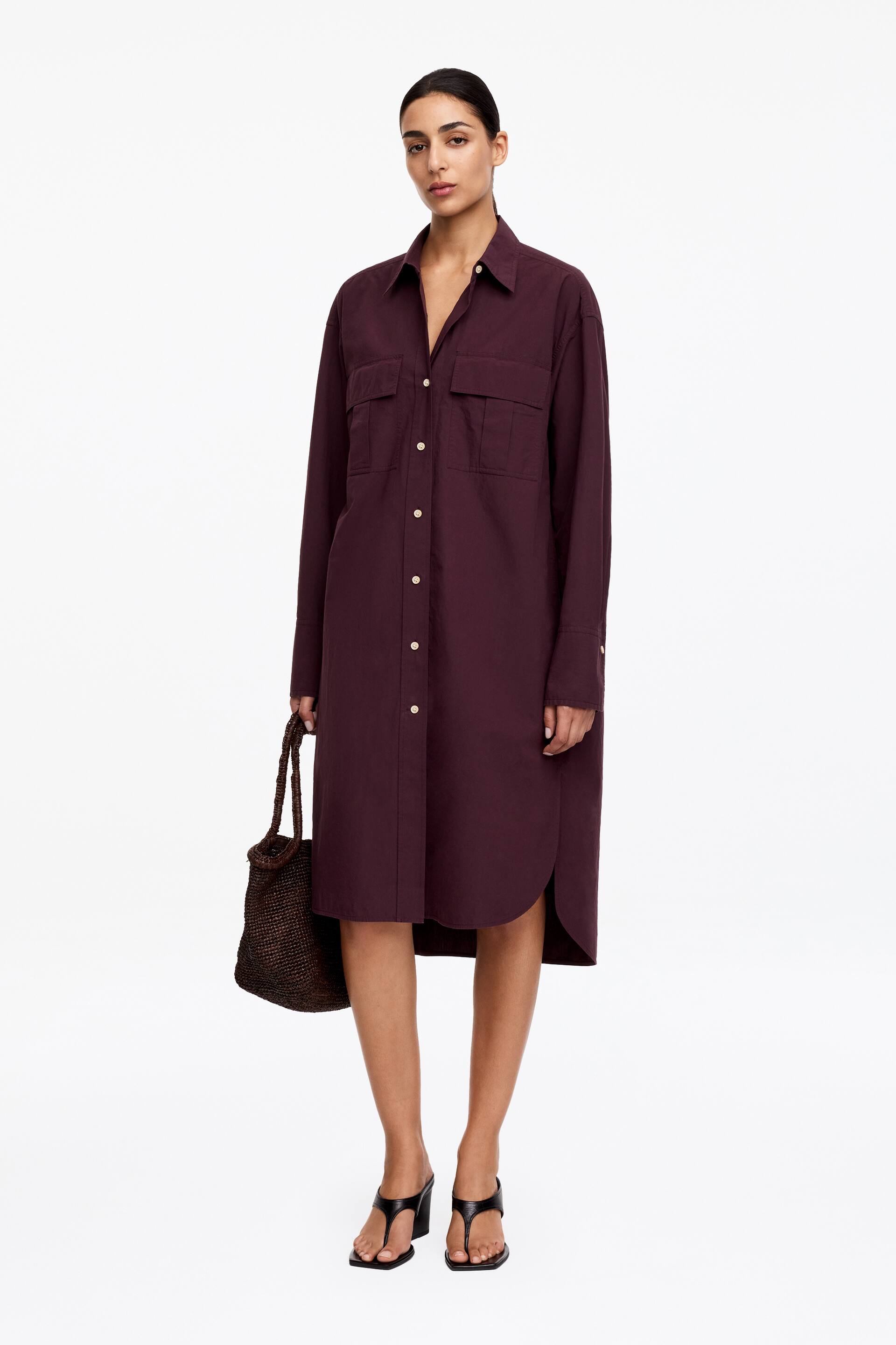 Agrandir l'image: Une robe-chemise prune foncé est présentée de face, avec un col à revers, des manches longues avec des poignets à bouton unique, et une patte de boutonnage complète avec des boutons de couleur claire. Deux poches à rabat sont visibles sur la poitrine, et la robe tombe juste en dessous des genoux avec des fentes arrondies sur les côtés.