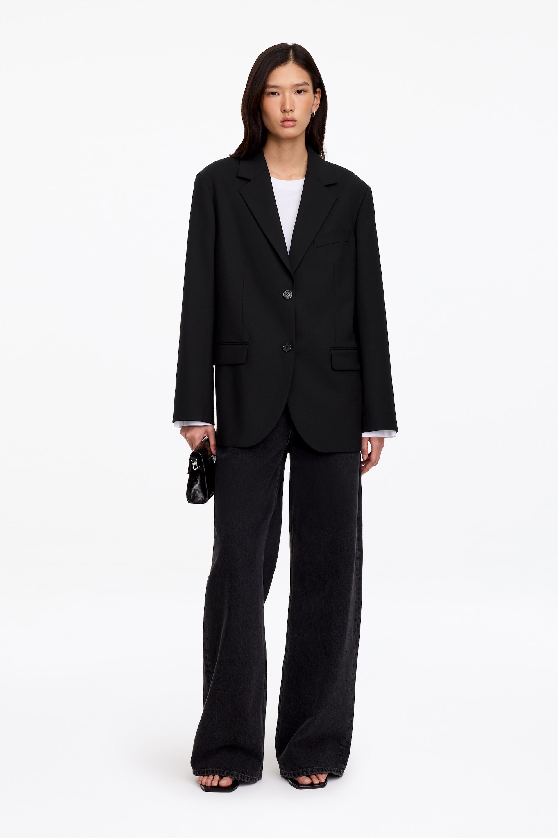 Vis større bilde: En svart blazer med avslappet passform og skreddersydd snitt bæres av modellen, lagvis over en hvit T-skjorte og kombinert med mørke jeans med vide ben. Den enkeltspente blazeren har spisse slag, frontlukking med to knapper og lommer med klaff, mens modellen holder en liten svart clutch i den ene hånden.