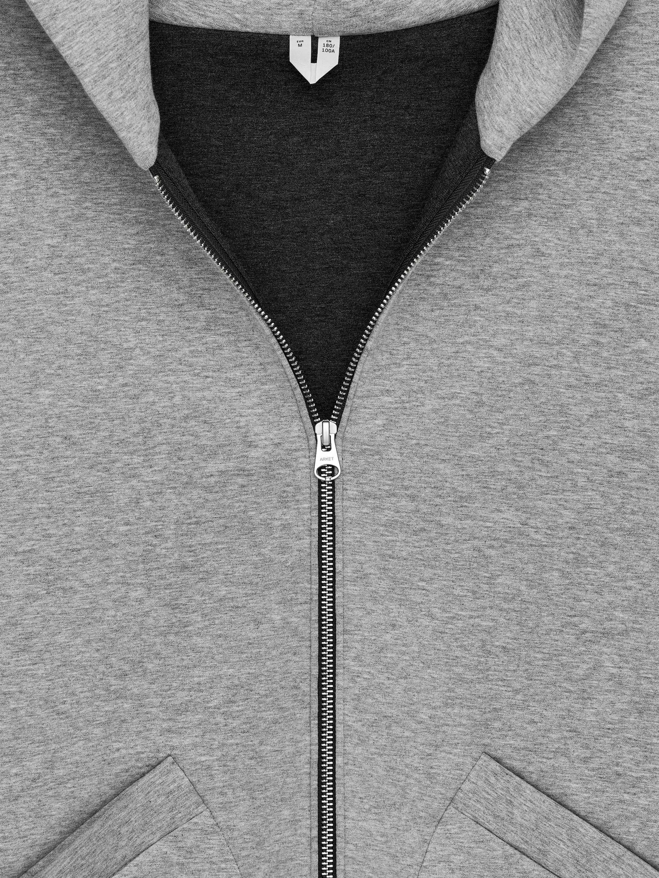 View larger image: A grey zipped hoodie is being presented from the front. It is featuring a partially unzipped central front zipper with a silver-toned pull. Slanted welt pockets are appearing at the bottom, and the inner hood is showing a dark grey color.