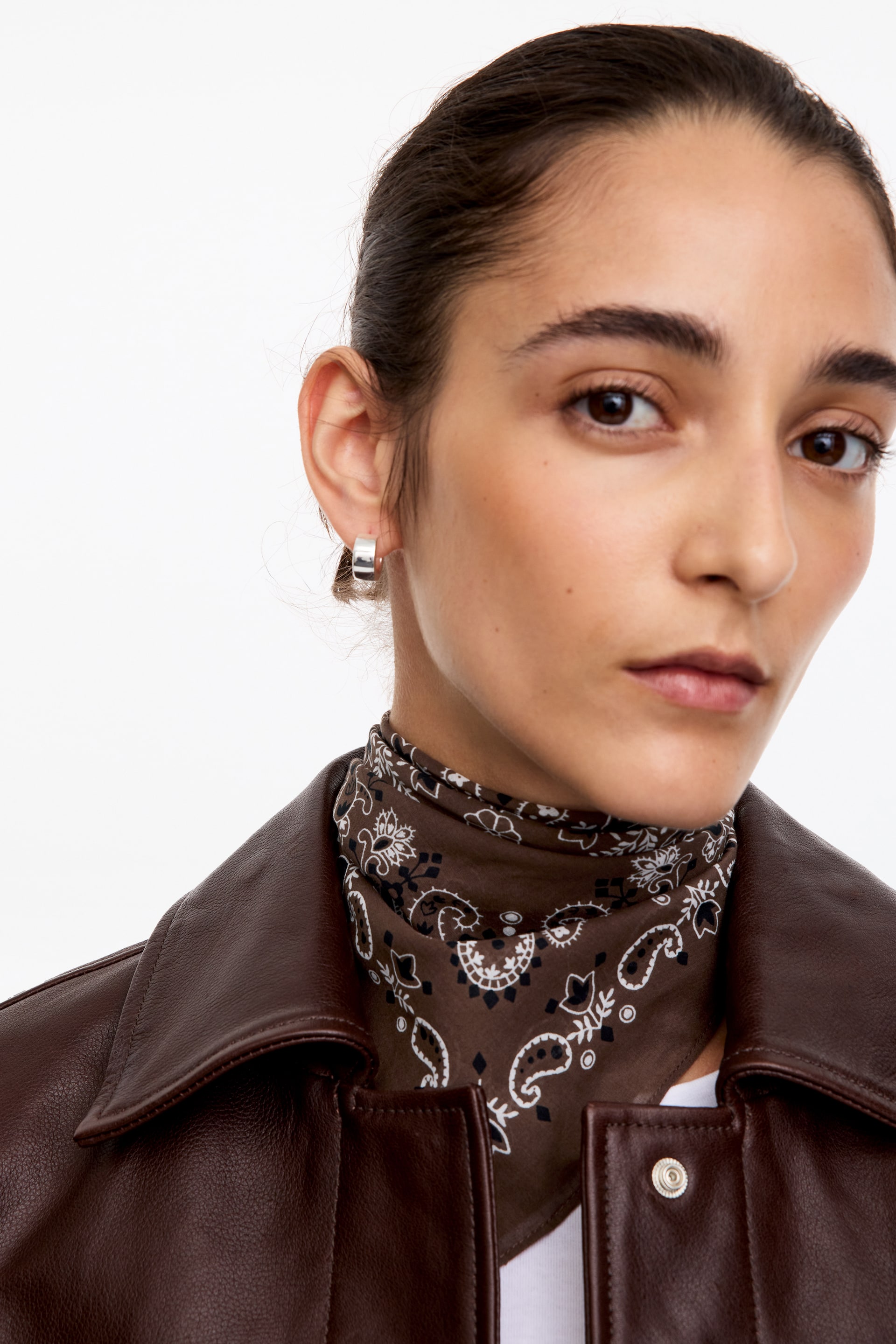 View larger image: A cotton scarf in brown with a white and black paisley pattern is being worn wrapped around the model's neck. The scarf is layered under a dark brown leather jacket, showcasing its detailed print.