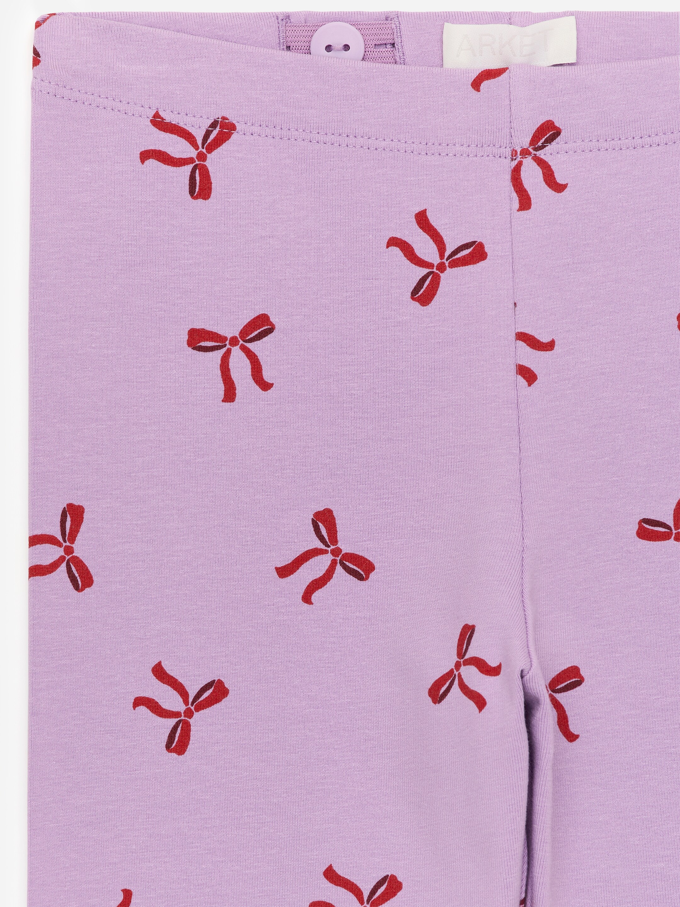 View larger image: Leggings are being presented in a light purple, soft single jersey fabric, patterned with small red bows. The front waistband is detailed with visible adjustable elastic and a button.