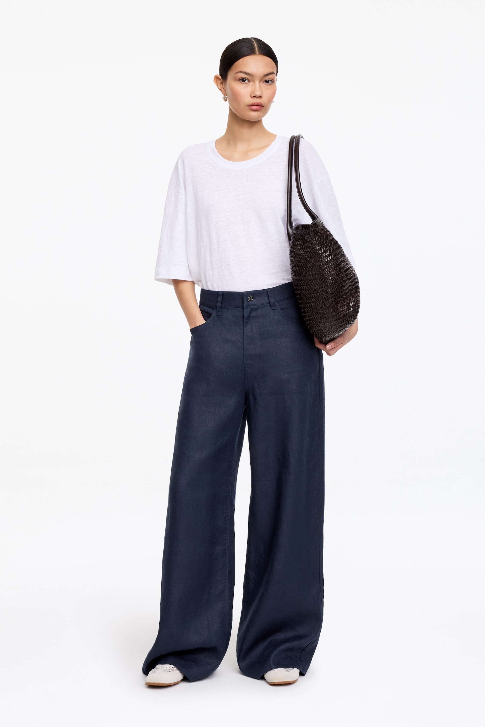 View larger image: Dark blue wide-leg trousers feature a high-waisted design with visible front pockets. The trousers exhibit a loose, flowing fit, with the full-length hems pooling slightly over light-coloured shoes.