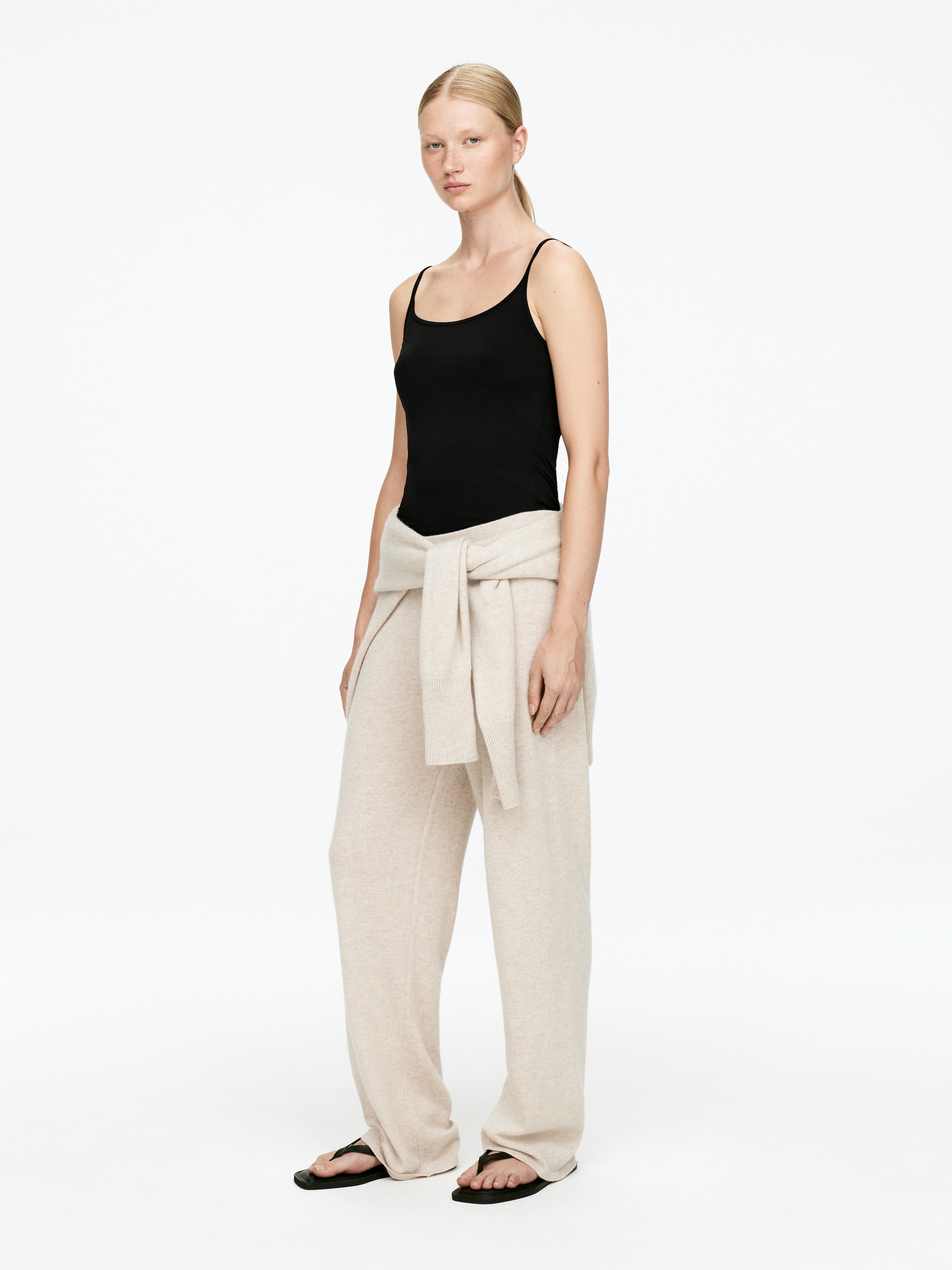 Visualizza immagine più grande: Un modello indossa pantaloni a maglia beige chiaro a gamba larga, in piedi principalmente rivolto in avanti, abbinati a un top nero e un maglione a maglia beige chiaro legato in vita.