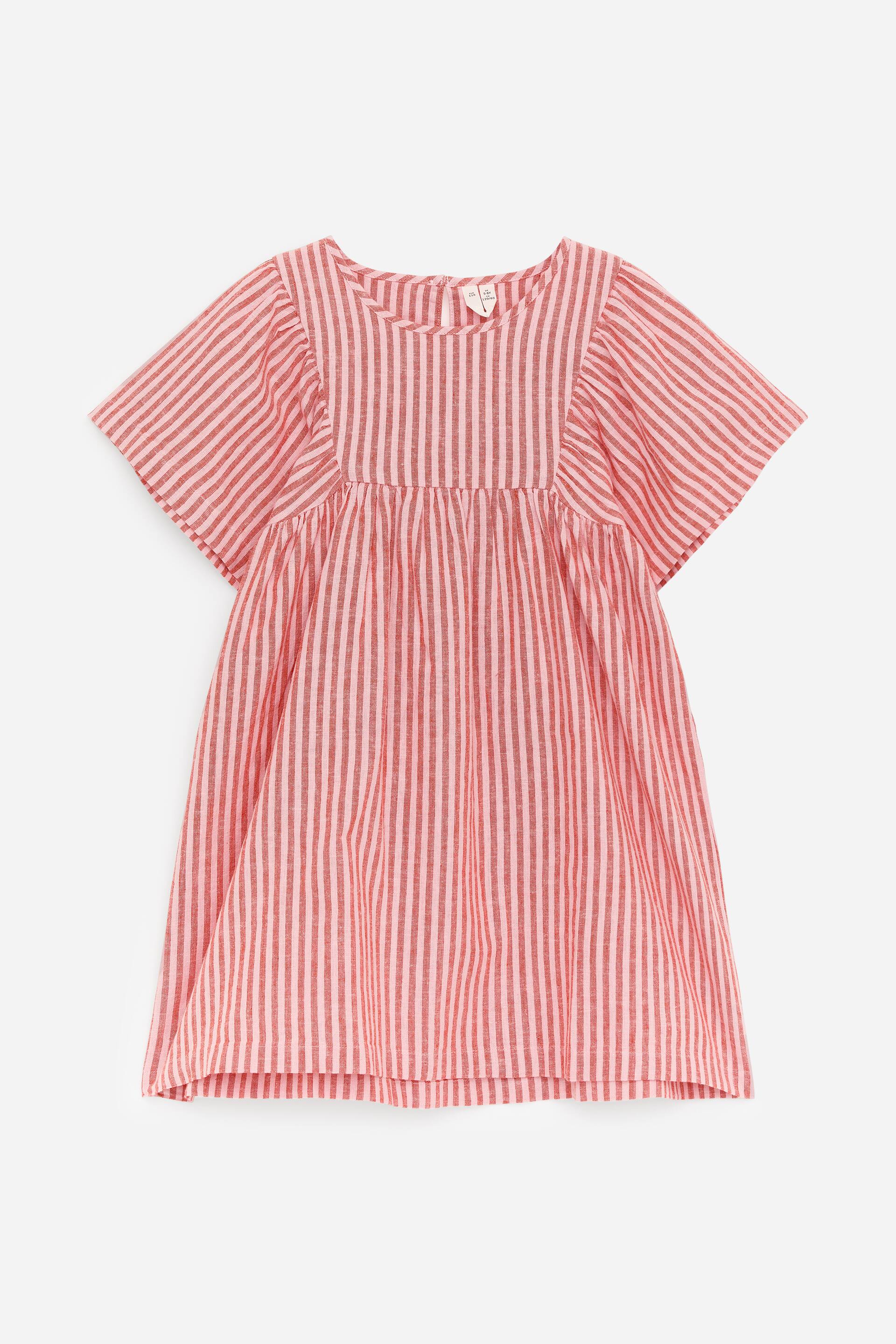Se større billede: Kjolen har en rund halsudskæring og korte flagrende ærmer, med en empire talje og rynker nedenunder. Den er mønstret med skiftende røde og lyserøde lodrette striber, der skaber en fyldig nederdel.