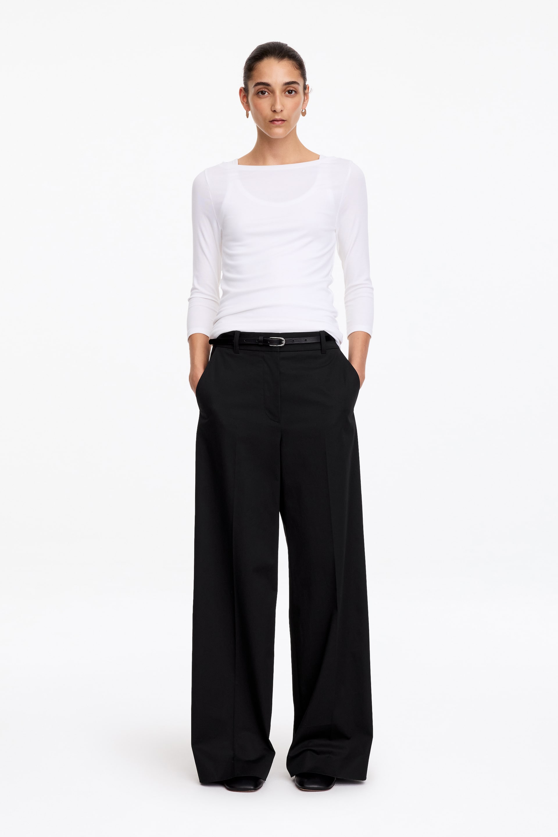 View larger image: Black high-waisted trousers featuring a straight-leg silhouette with pressed creases and side pockets are worn by the model. The model is standing with hands in the pockets, styling the trousers with a slim black belt and a white long-sleeved top.