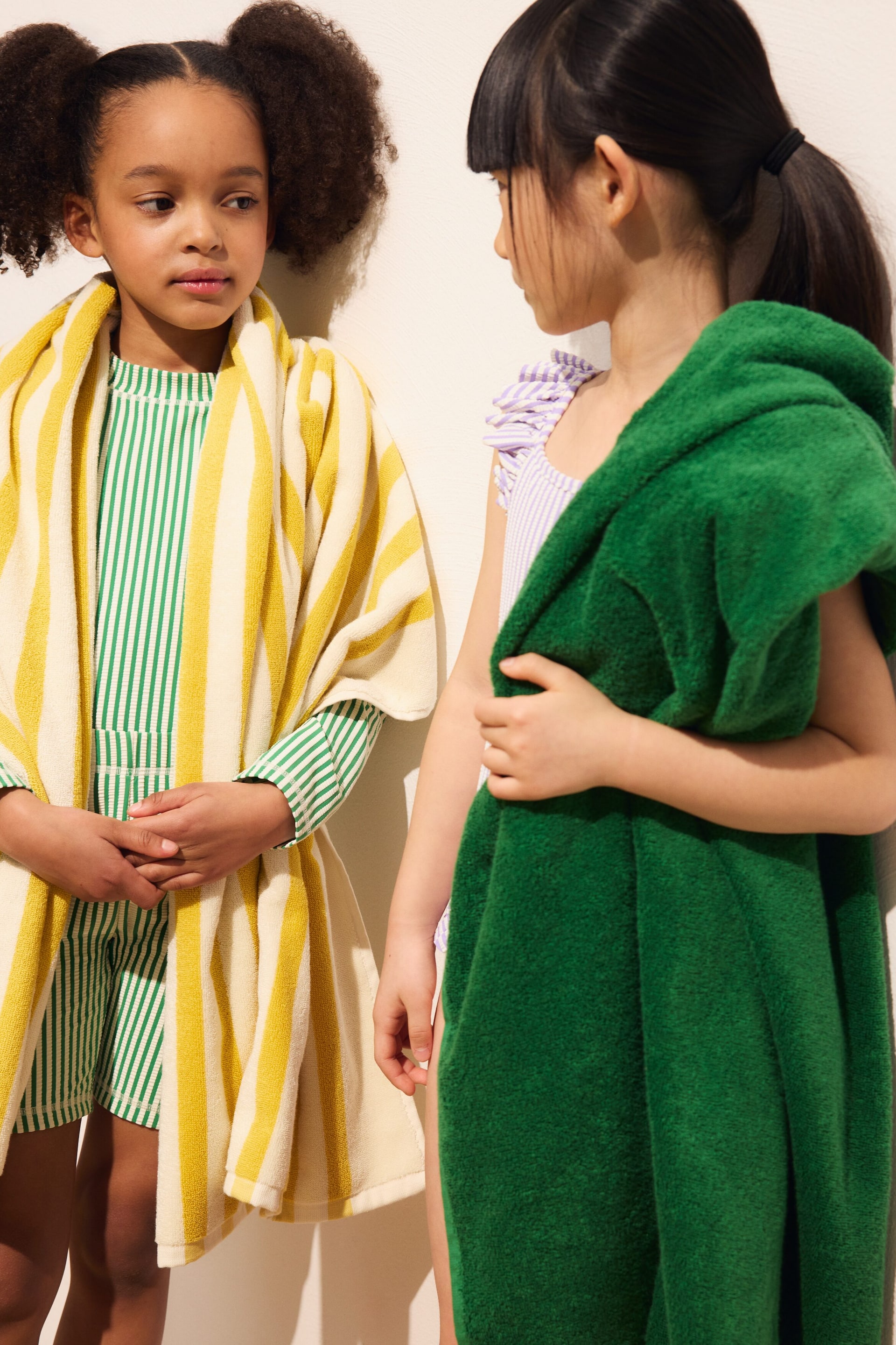 Größeres Bild anzeigen: Ein gelb-weiß gestreiftes Handtuch-Poncho ist über die Schultern drapiert, mit breiten vertikalen Streifen und einer flauschigen Frottee-Textur, die ein grün-weiß gestreiftes Oberteil und Shorts bedeckt. Ein dunkelgrünes Handtuch wird über der rechten Schulter gehalten, ebenfalls aus Frottee gefertigt.