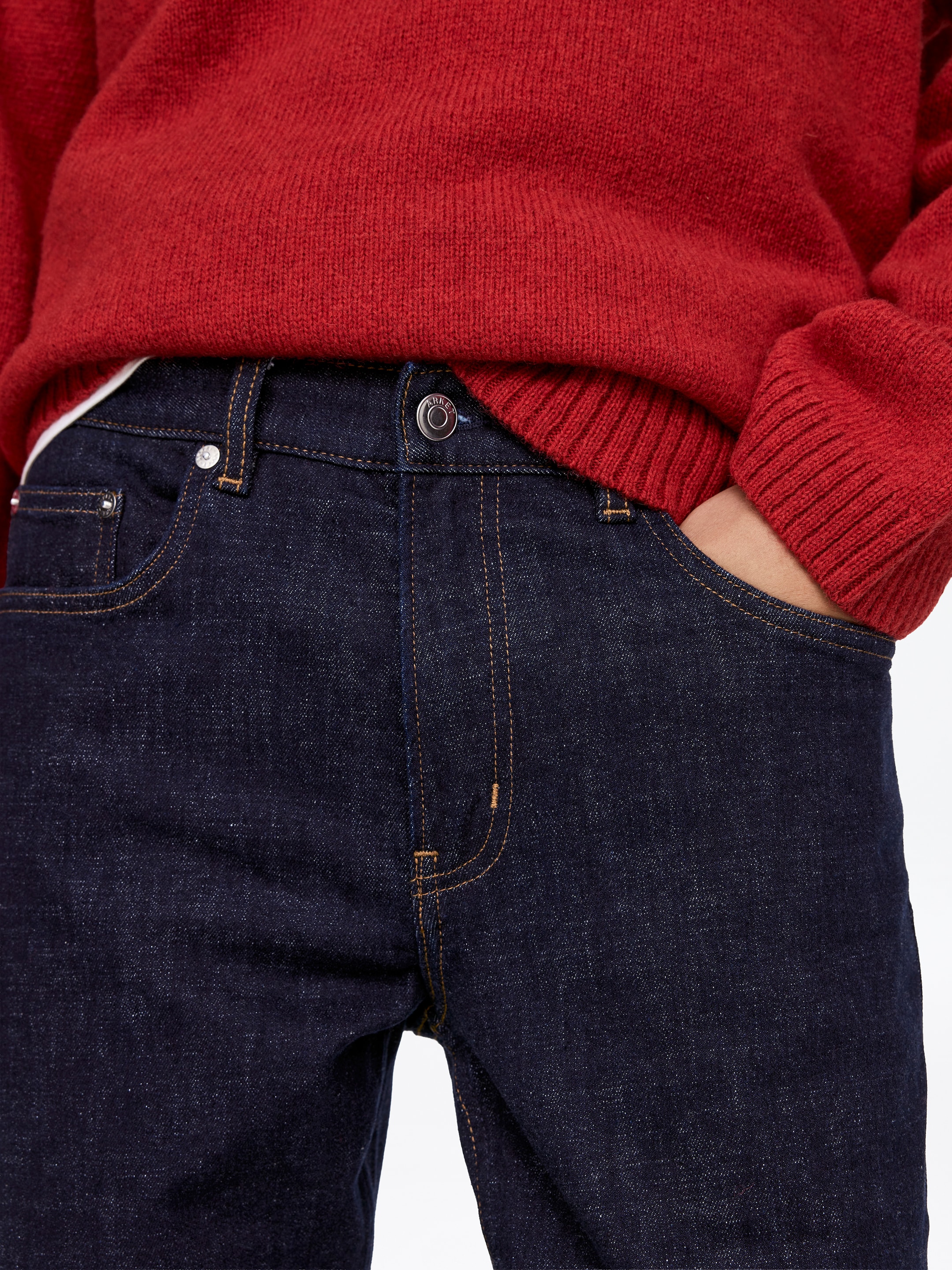Größeres Bild anzeigen: Dunkel indigoblaue Denim-Jeans werden aus einer frontalen, leicht angewinkelten Perspektive präsentiert, wobei der Fokus auf der Taille und dem oberen Oberschenkel liegt. Sie zeichnen sich durch kontrastierende hellbraune Nähte, einen Metallknopfverschluss am Bund und Metallnieten an den Vordertaschen aus, während ein roter Strickpullover teilweise eingesteckt ist, was auf die Passform hinweist.