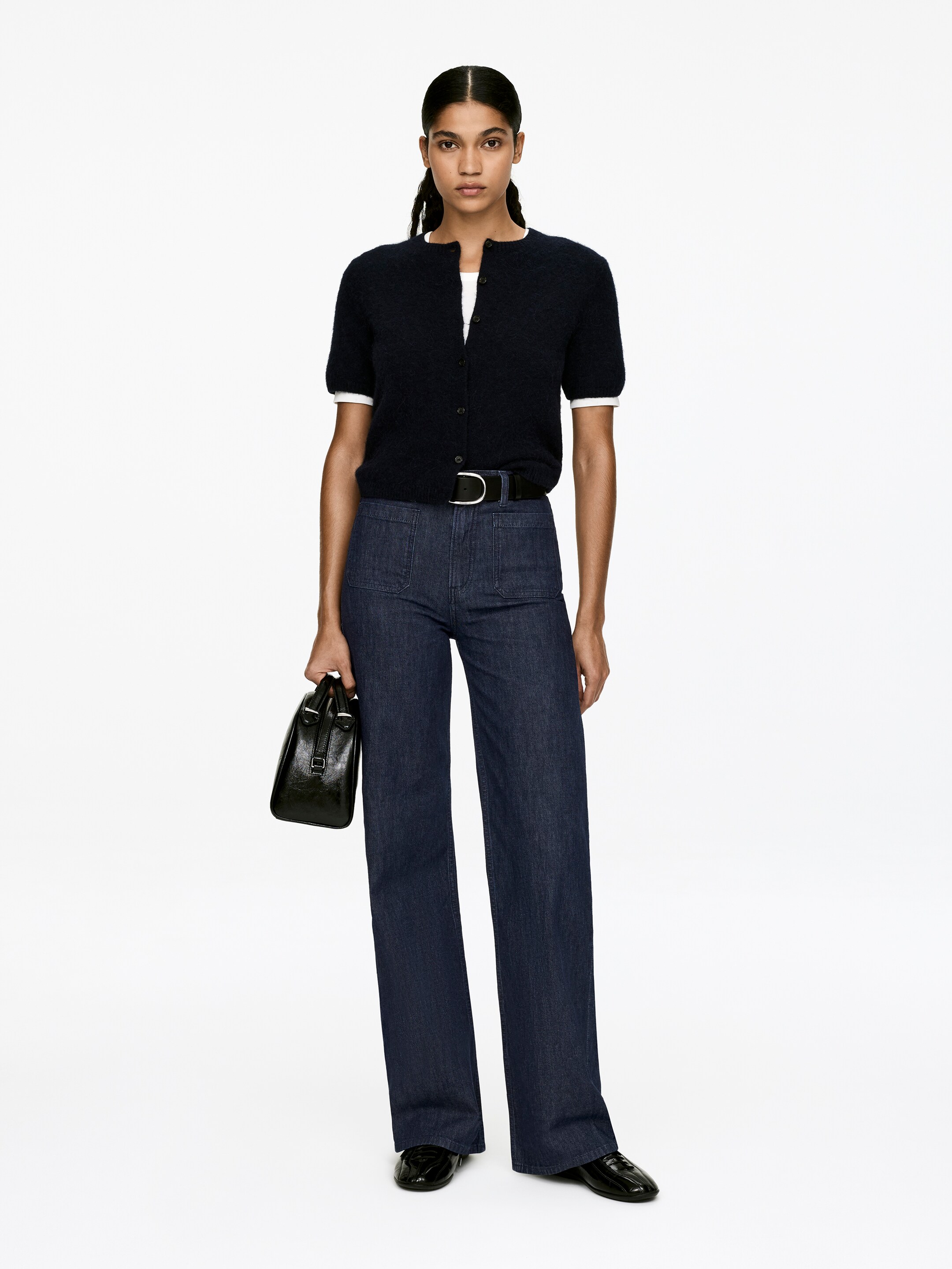 Agrandir l'image: Une tenue est présentée, face à l'avant. Un cardigan à manches courtes, texturé moelleux et bleu marine foncé, superposé sur un t-shirt blanc à col rond, est associé à un jean à taille haute et à jambes larges, avec une teinte foncée, doté de poches avant et d'une ceinture noire avec une boucle de ton argenté. Des mocassins en cuir verni noir sont visibles et un sac à main structuré noir est porté.