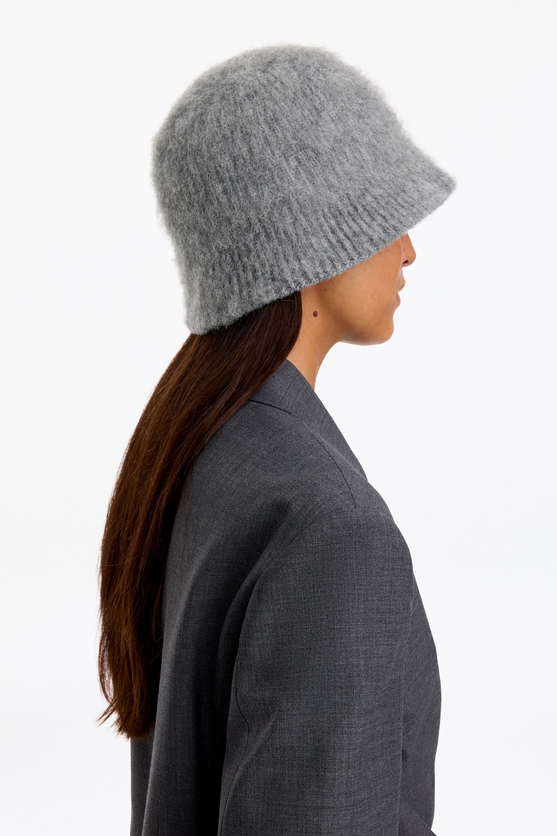 Zobraziť väčší obrázok: Svetlosivý bucket hat z brúseného úpletu s oblým vrchom a jemne rozšíreným okrajom má na sebe model. Model je zobrazený v profile, pričom klobúk je štylizovaný s tmavosivým štruktúrovaným sakom.