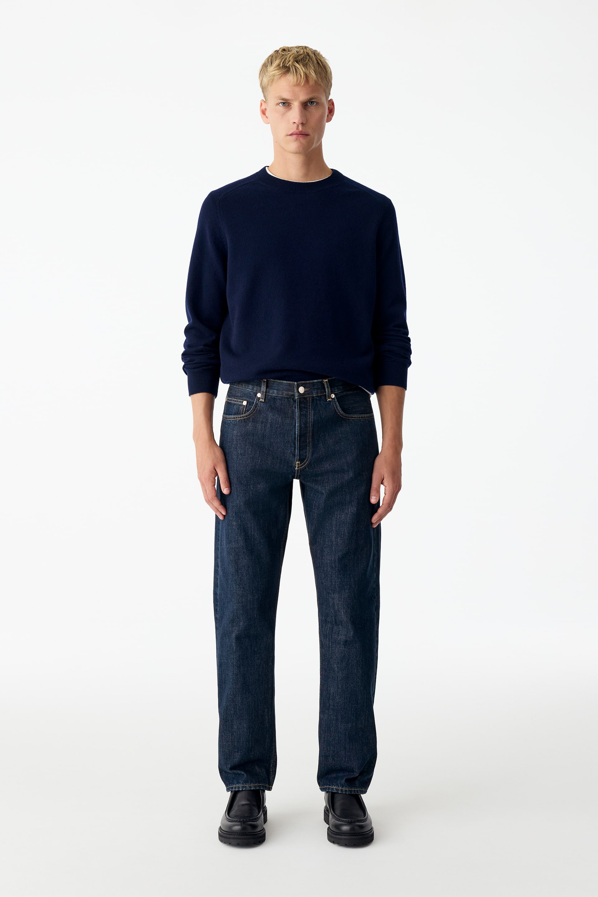 Größeres Bild anzeigen: Diese gerade geschnittene blaue Denim-Jeans in dunkler Waschung wird vom Model getragen und zeichnet sich durch einen mittelhohen Bund sowie eine lockere Passform in voller Länge aus. Die Jeans ist mit einem in den Bund gesteckten marineblauen Rundhalspullover und schwarzen Lederschuhen kombiniert.