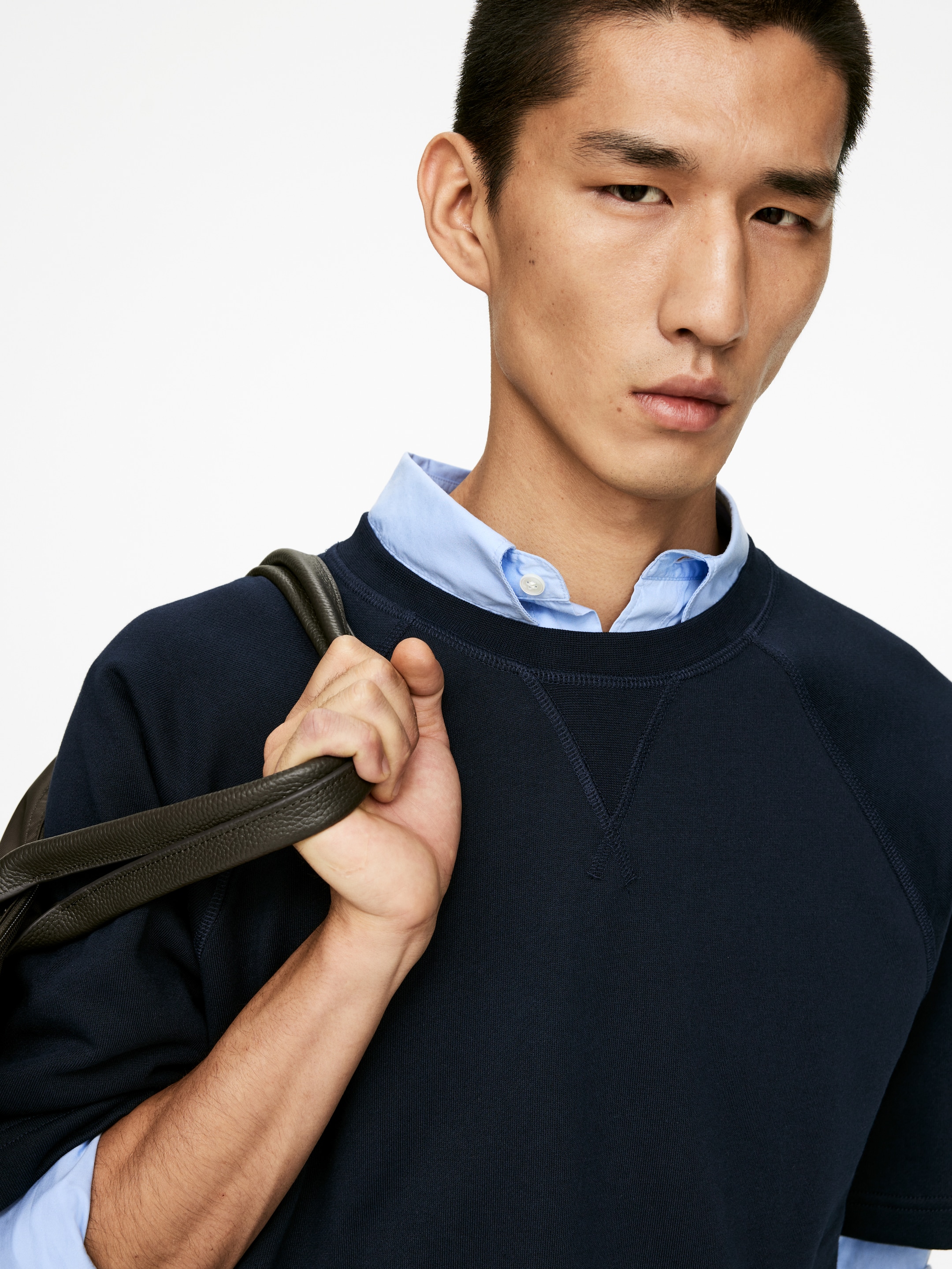 Größeres Bild anzeigen: Ein marineblaues Sweatshirt mit Rundhalsausschnitt wird aus einem Viertelwinkel von vorne präsentiert, wobei das V-Stich-Detail unter dem Kragen und die Raglanärmel zur Geltung kommen. Ein hellblaues Hemd mit Kragen ist darunter geschichtet und schaut am Halsausschnitt hervor.