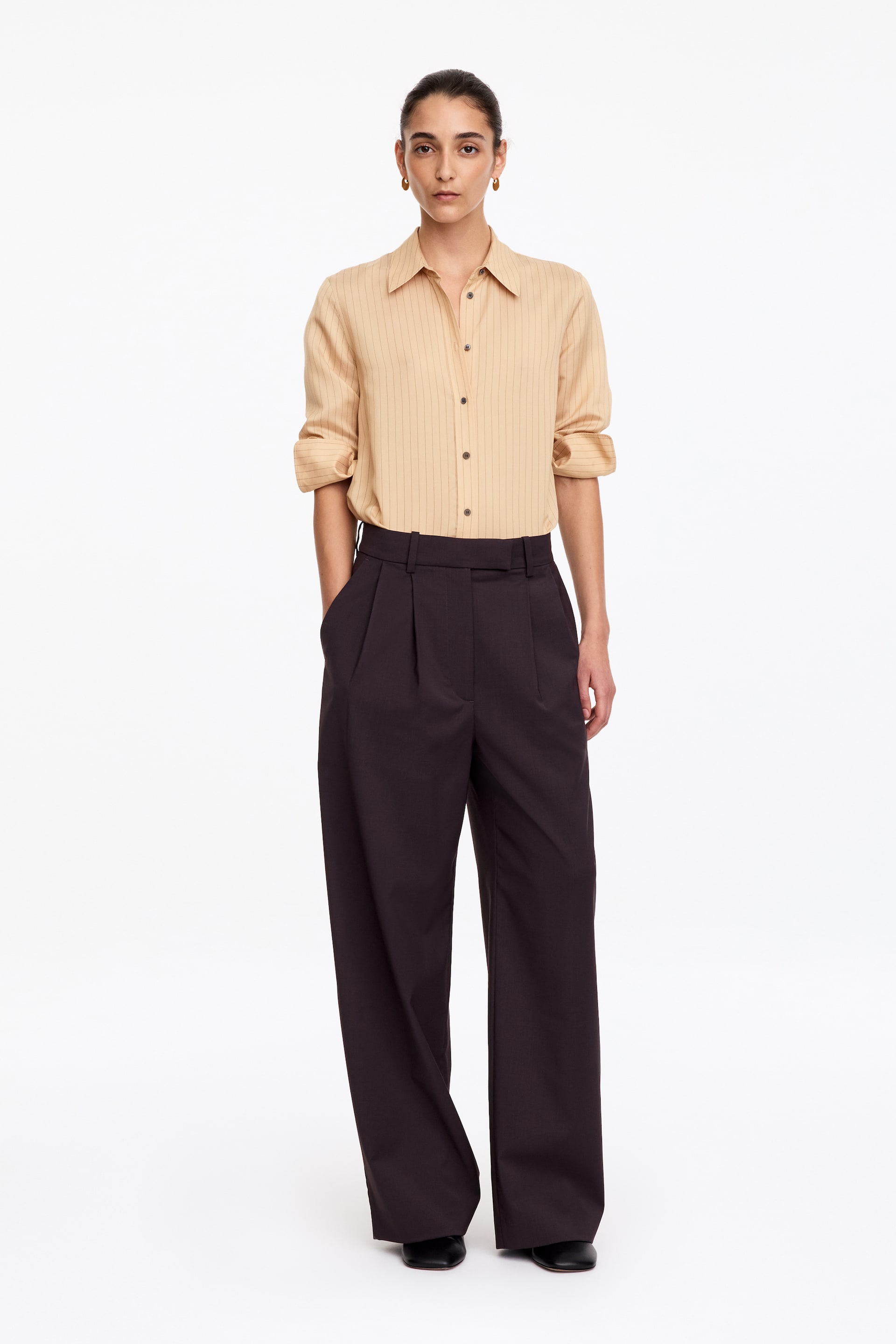 Grotere afbeelding bekijken: Deze donkere wide-leg broek, met een hoge taille en plooien aan de voorkant, wordt gedragen door het model. Het model staat met één hand in de zijzak en stylet de broek met een ingestopte lichtbeige gestreepte blouse.