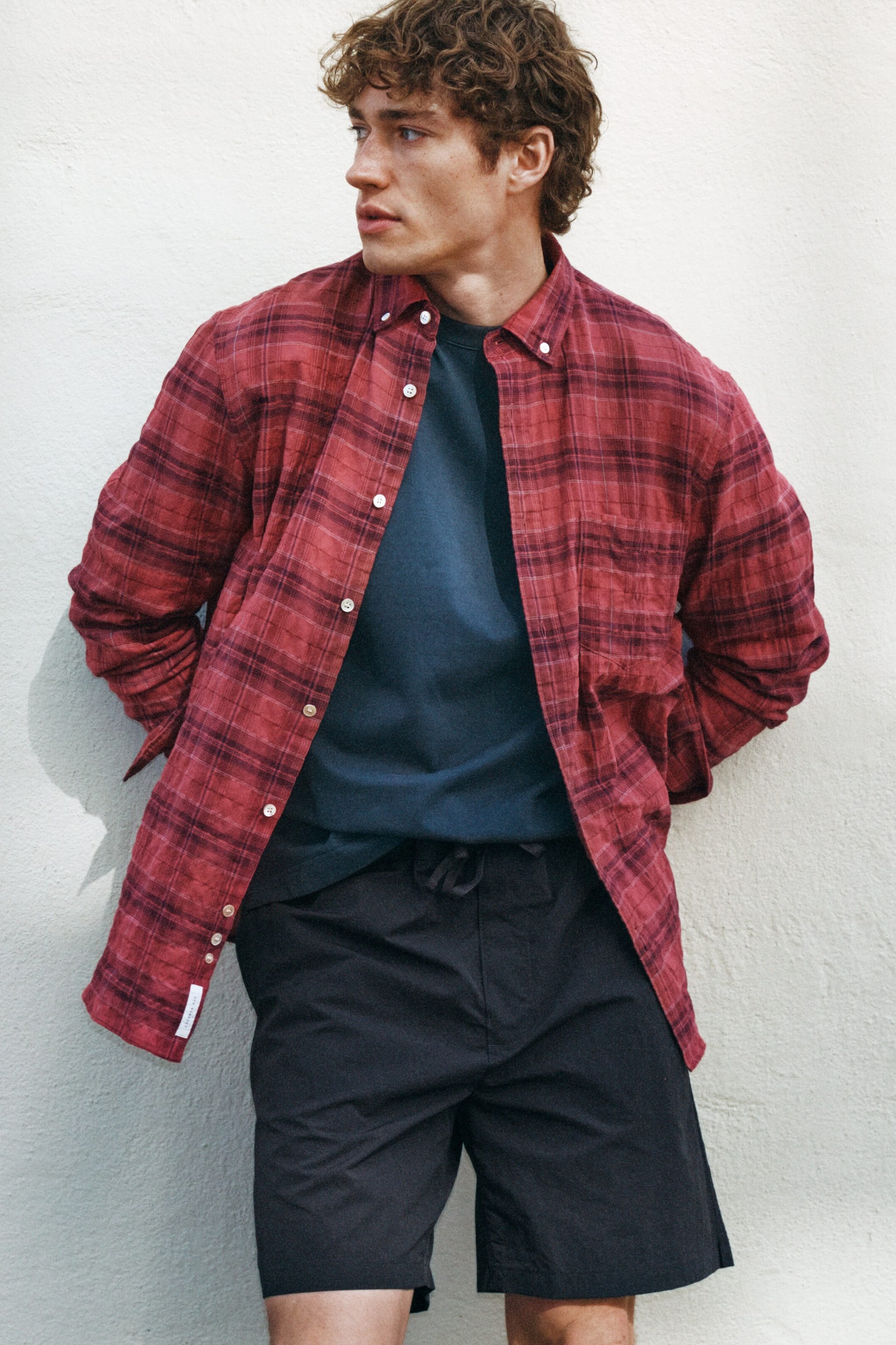 Größeres Bild ansehen: Ein rotes, kariertes Button-Down-Hemd wird offen über einem dunkelblauen Rundhals-T-Shirt getragen. Es verfügt über ein rot-dunkelrotes Karomuster, einen Button-Down-Kragen mit weißen Knöpfen und eine sichtbare Brusttasche.