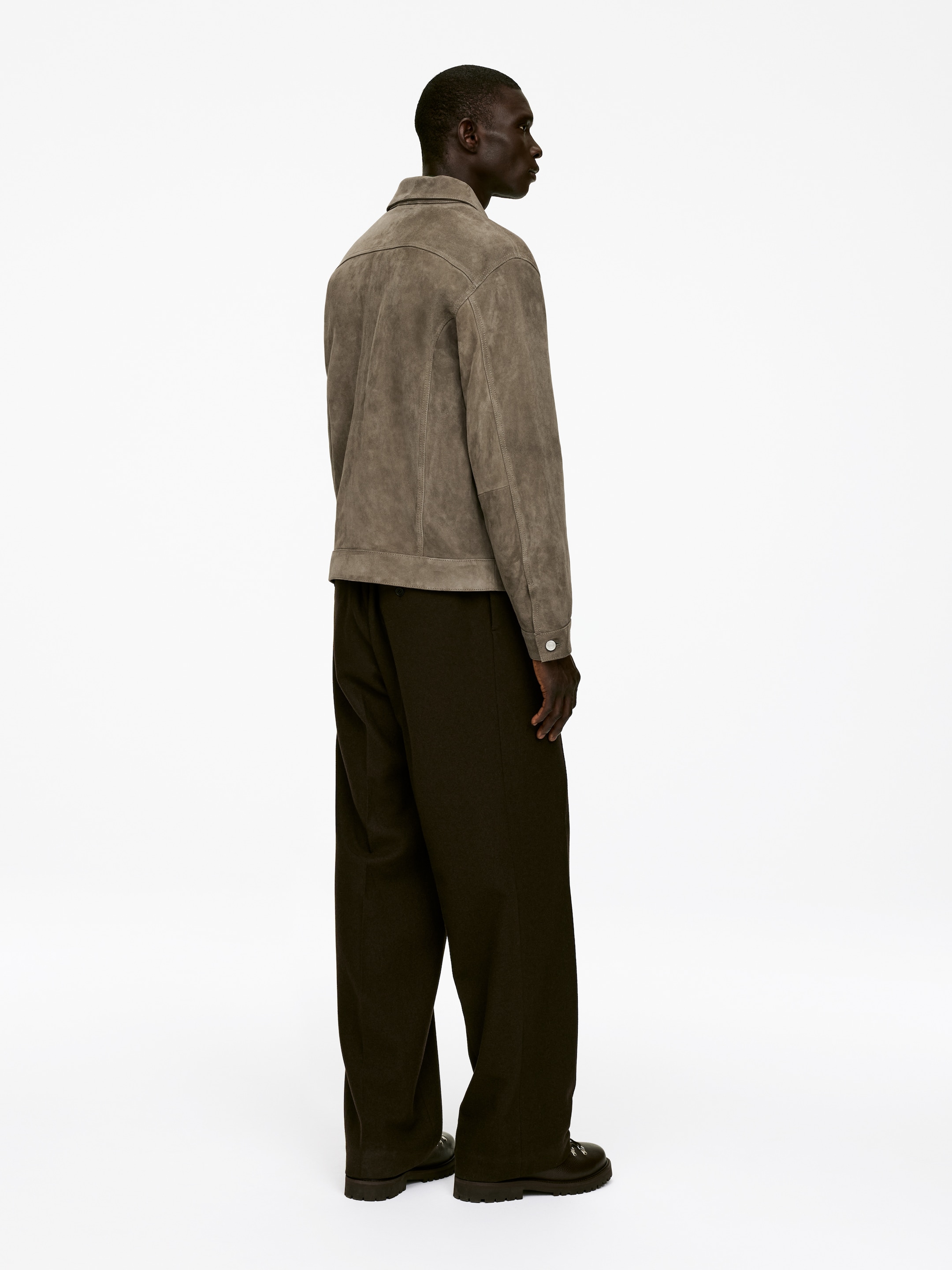 Agrandir l'image: Une veste en daim brun-grisâtre est portée, présentée sous un angle arrière-latéral, mettant en valeur sa coupe décontractée, son col classique et ses coutures de panneaux visibles à travers le dos. Elle est associée à un pantalon large et sombre et des chaussures sombres à semelles crantées.