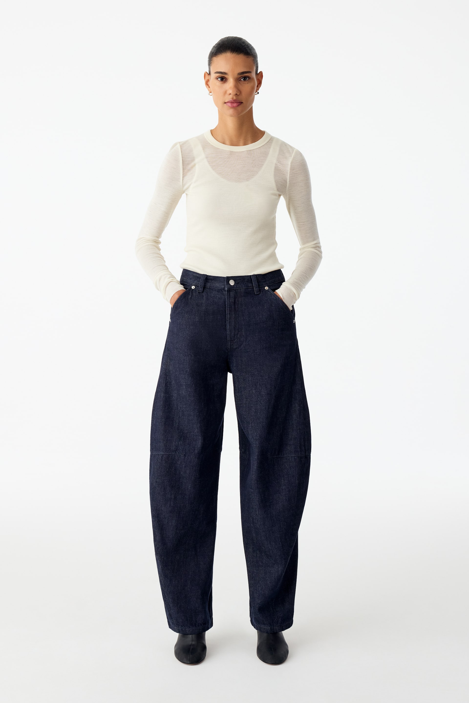 Visualizza immagine più grande: Jeans in denim scuro con vita media e silhouette rilassata a gamba barrel, indossati dalla modella che è in piedi con le mani nelle tasche anteriori. I jeans sono abbinati a un top crema a maniche lunghe infilato dentro e a scarpe nere.