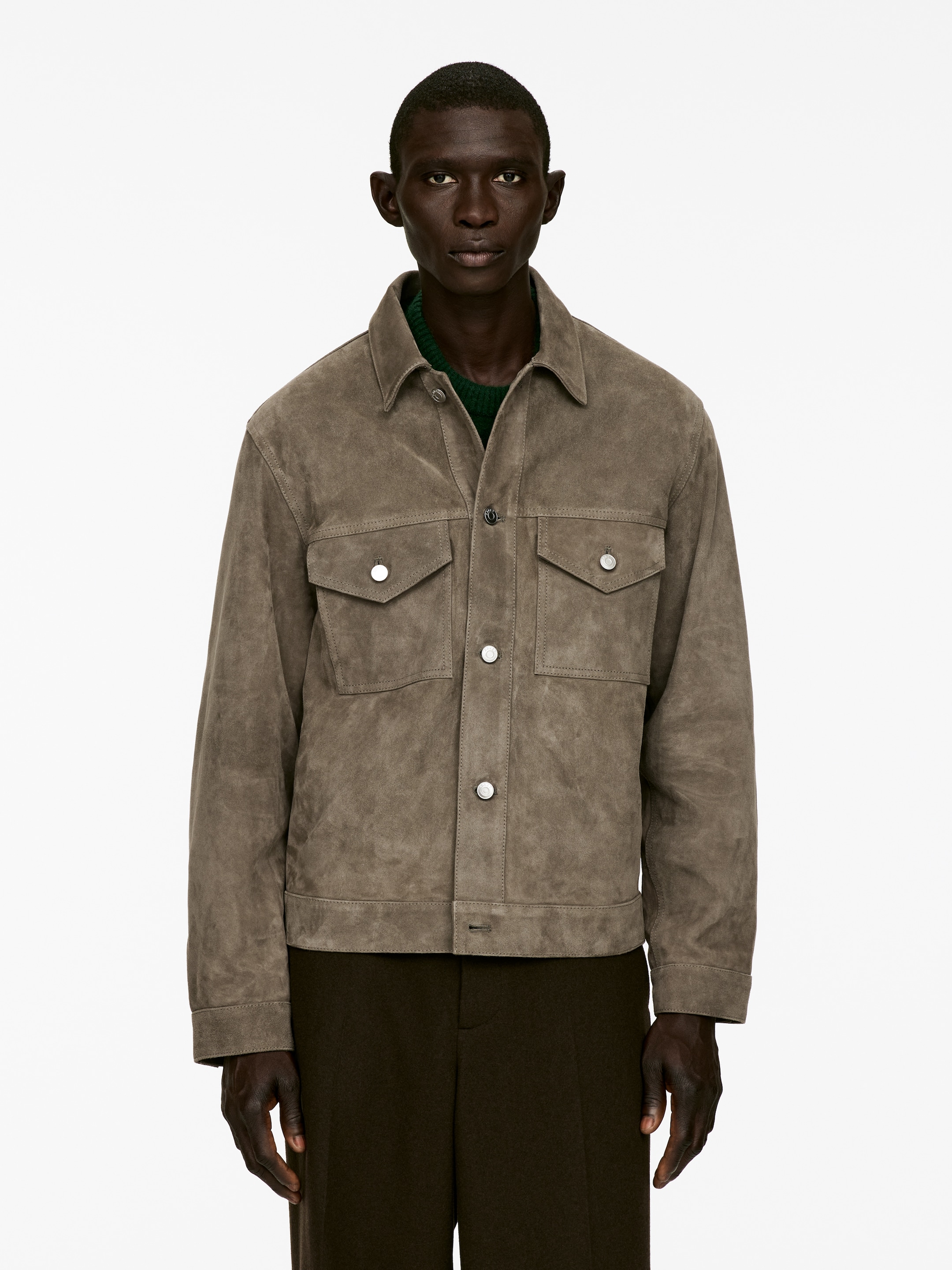 Agrandir l'image: Une veste-chemise en daim taupe est présentée de face, avec un col classique en pointe, une fermeture complète à boutons-pression et deux poches à rabat sur la poitrine, chacune fermée par un bouton-pression argenté, et est superposée sur un pull en maille vert.