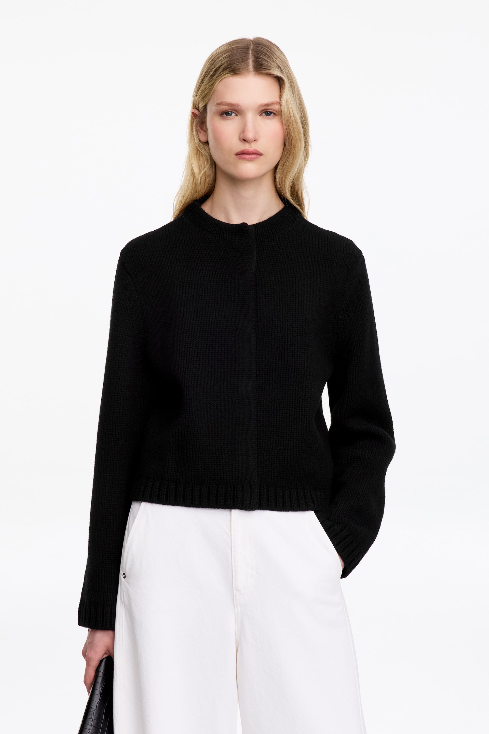 Ver imagen más grande: La modelo lleva un cárdigan de lana negro con cuello alto simulado, bajo de canalé y tapeta frontal oculta. El cárdigan se combina con pantalones blancos de pierna ancha, mientras la modelo mete una mano en el bolsillo del pantalón y sostiene un clutch negro texturizado.