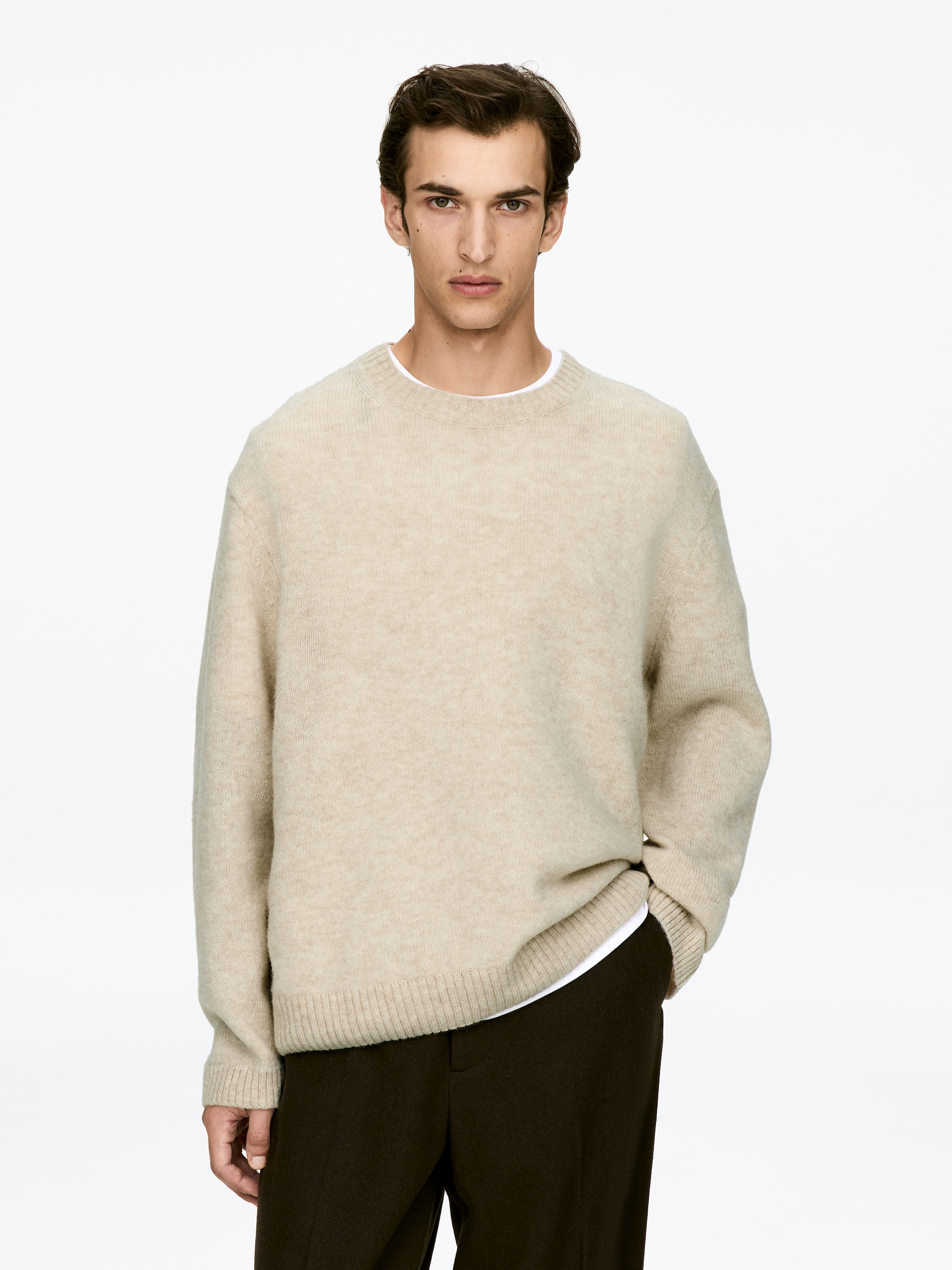 Visa större bild: En beige tröja med rund hals är vänd framåt, med en avslappnad passform och en mjuk, borstad stickad textur. Den visar upp ribbade muddar och en ribbad fåll, med en vit t-shirt som subtilt lager under.