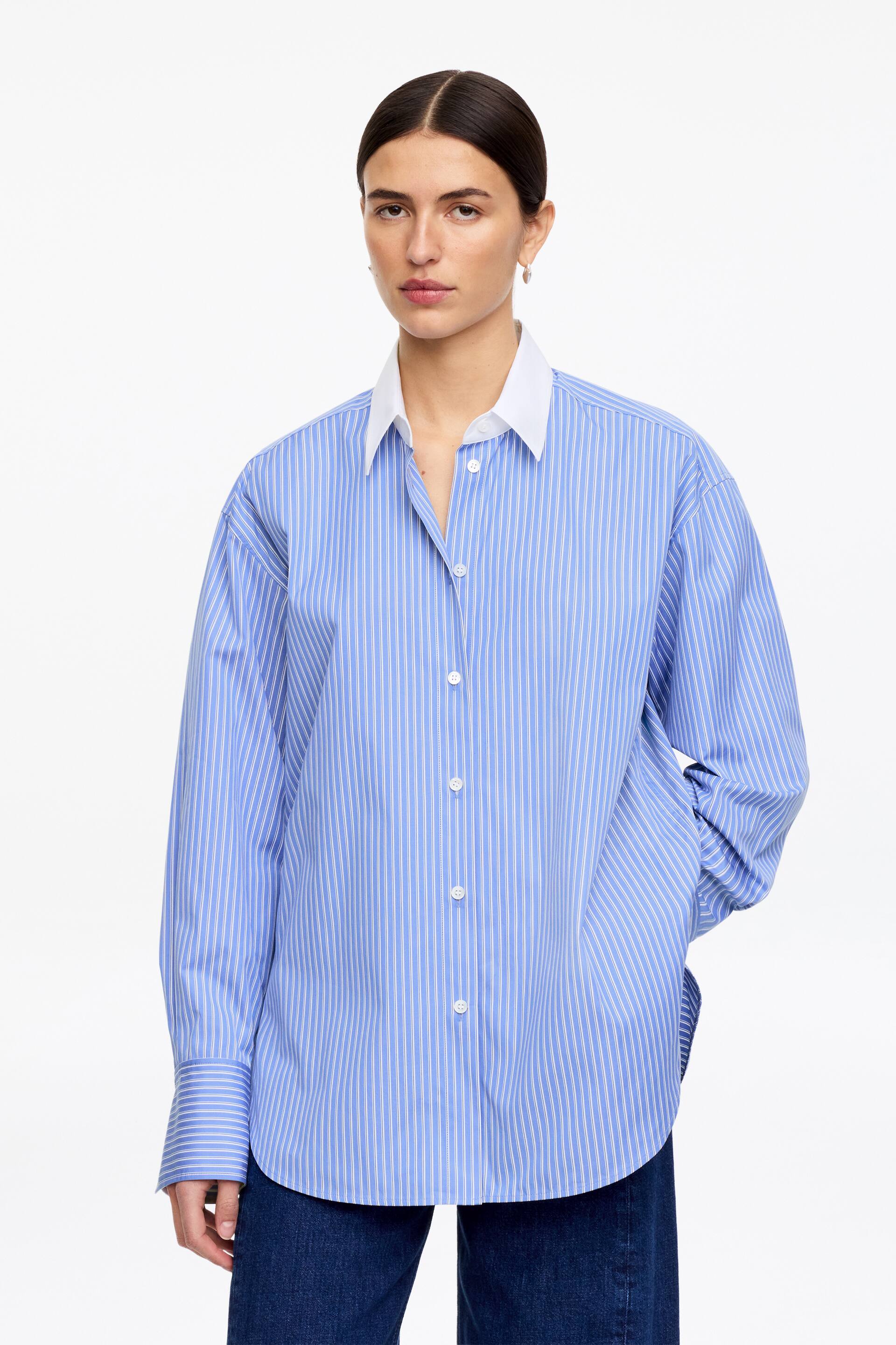 Se større billede: En blå- og hvidstribet skjorte med knapper vender fremad og har en kontrasterende hvid spredkrave, en central placket med hvide knapper og lange, løse ærmer, der afsluttes med knappede manchetter. En afrundet kant fuldender skjortens nederste kant.